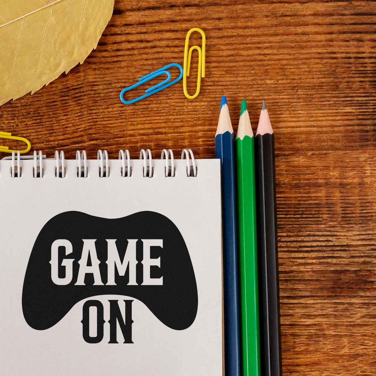 Game On stamp and seal design on notebook, with black controller graphic, blue, green and black pencils on a wood desk.