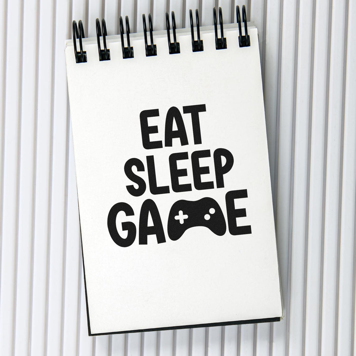 Black spiral-bound stamp design with EAT SLEEP GAME text and game controller icon on a white pad