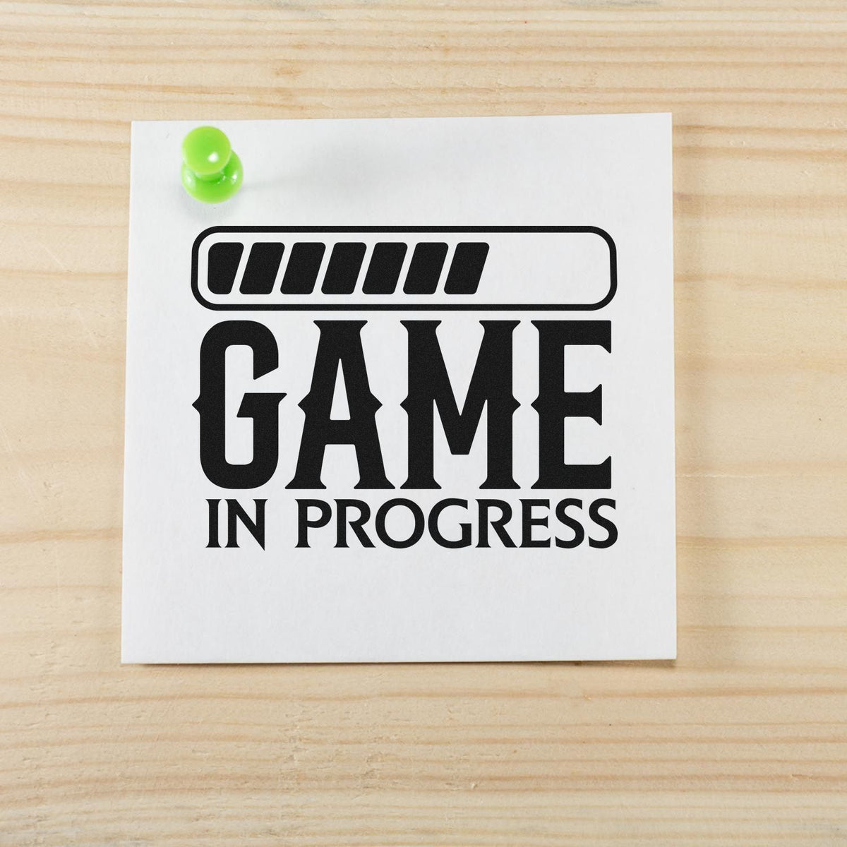 Black Game in Progress stamp and seal design on white paper, pinned with a bright green tack on light wood.