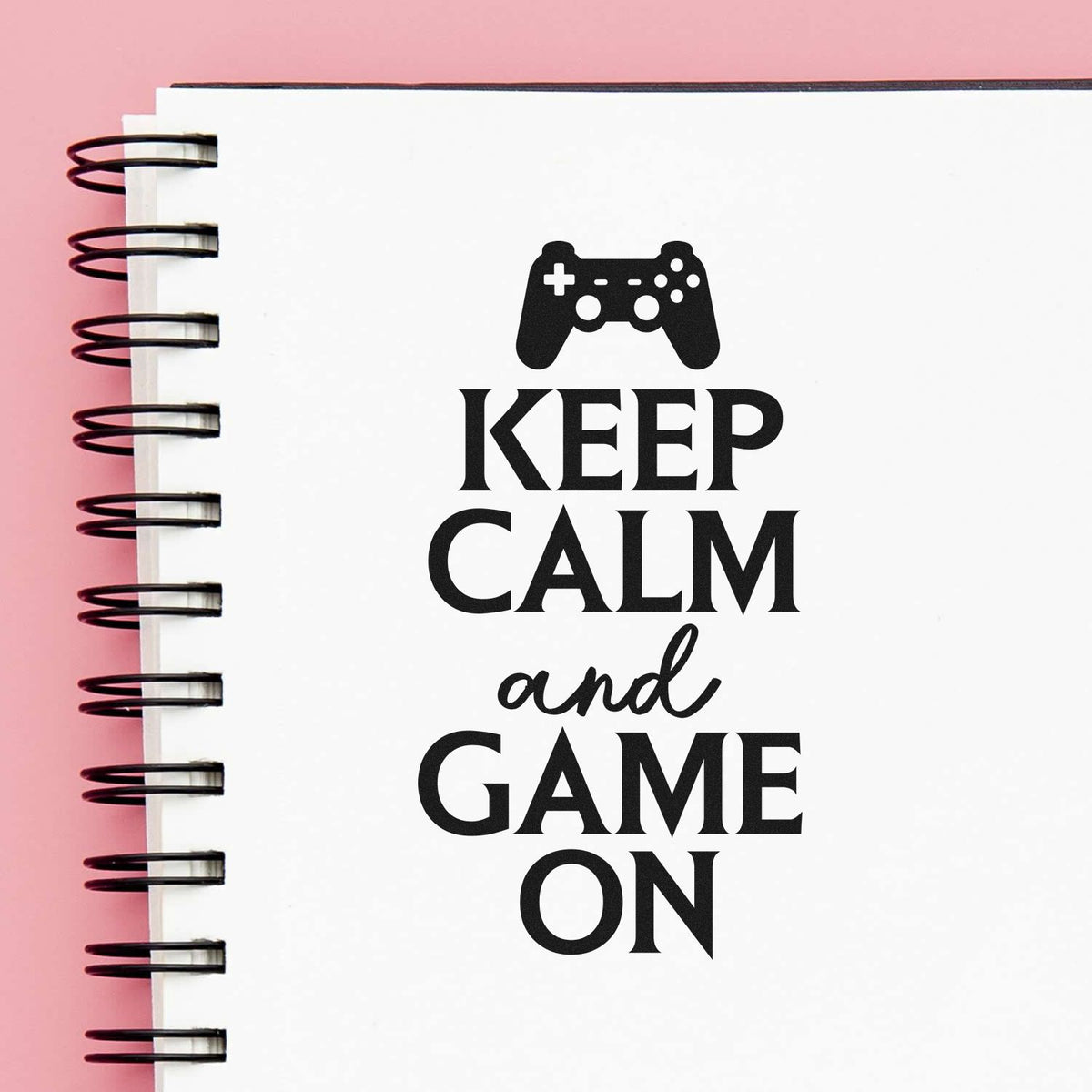 Black game controller stamp and seal reading Keep calm and game on on a white spiral notebook with pink background.