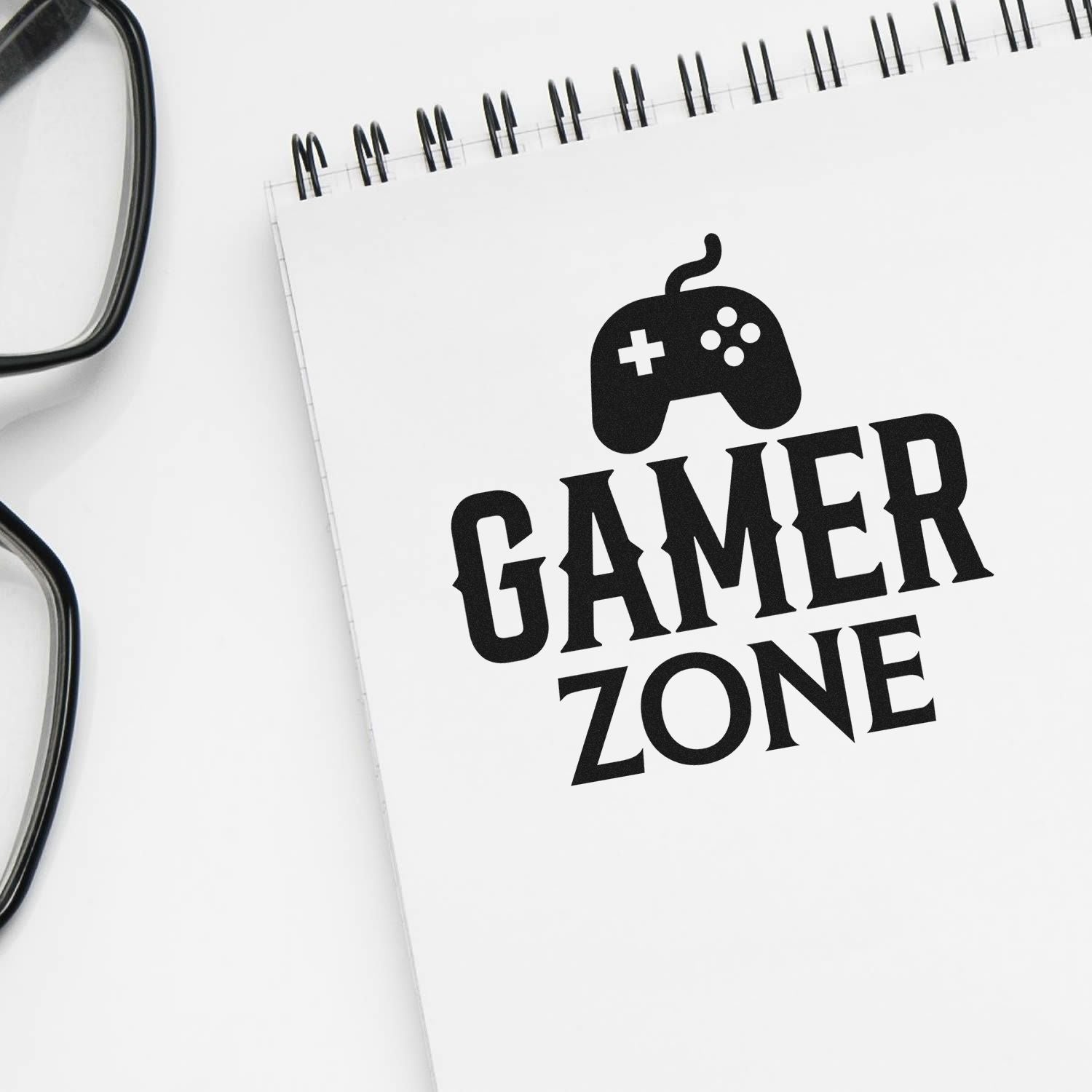 Black Gamer Zone stamp seal with game controller icon on white spiral notebook, beside black eyeglasses.