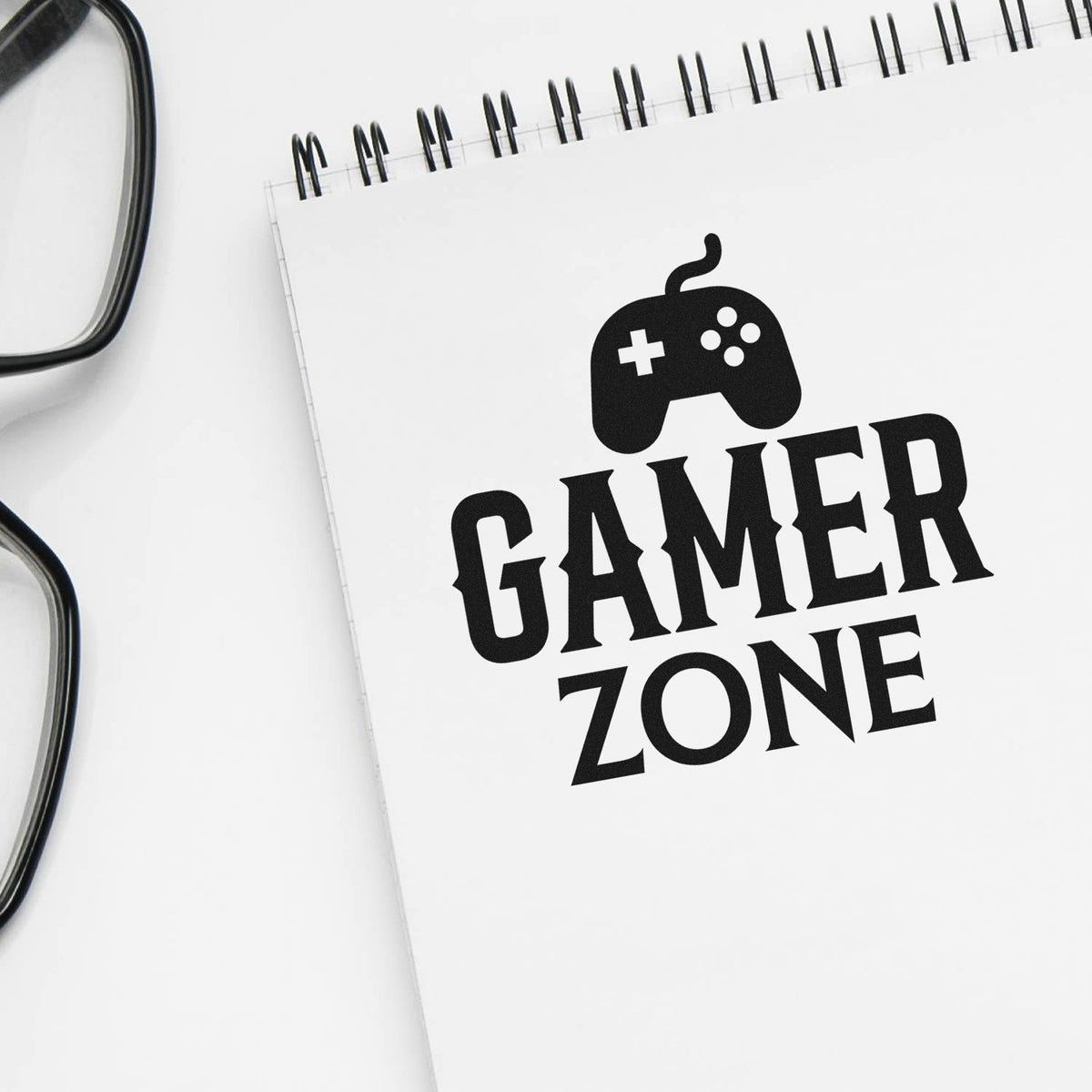 Black Gamer Zone stamp and seal design on white spiral notebook, with black game controller icon and bold lettering.