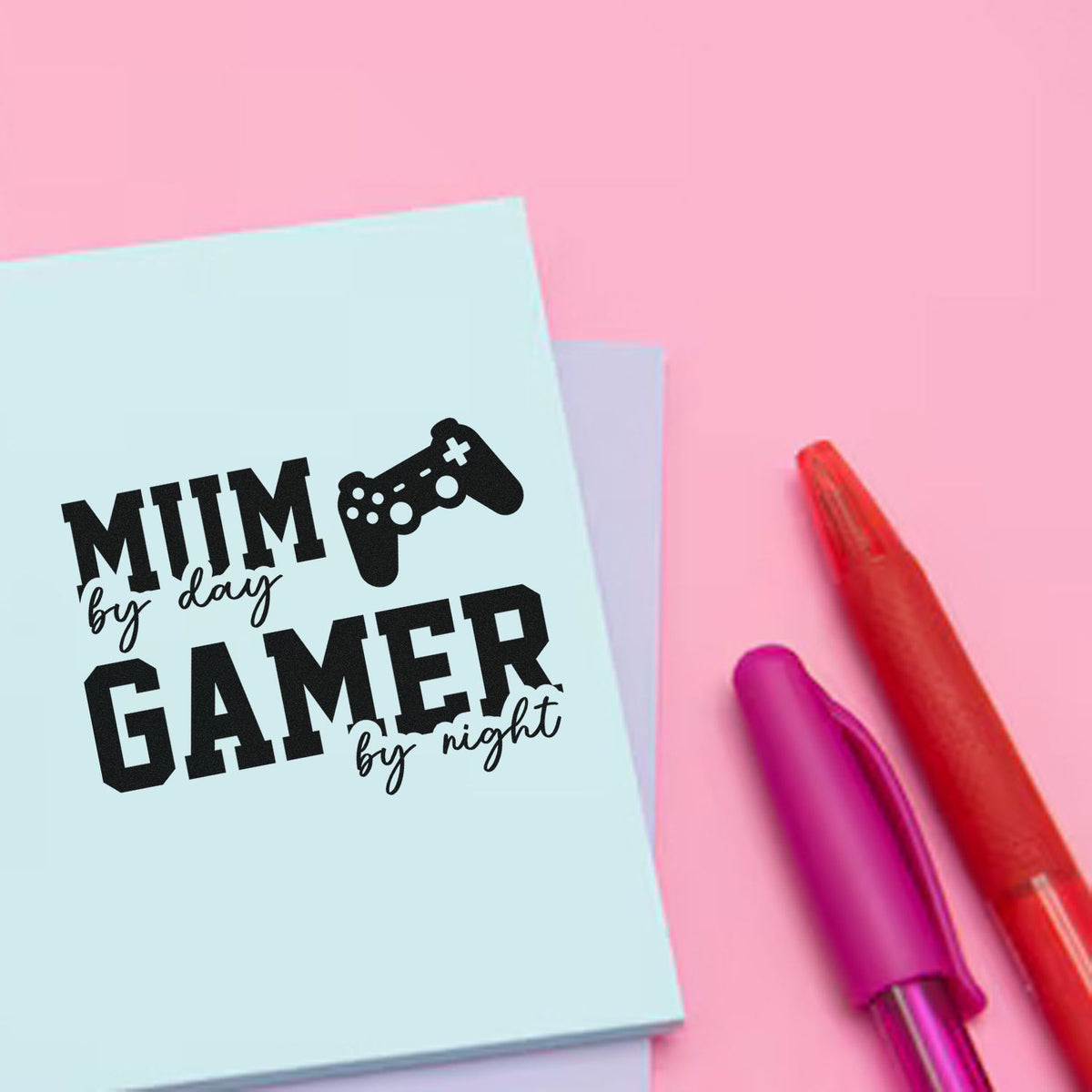 Light blue stamp seal with black Mum by day, gamer by night text and game controller on pink background, red and purple pens.