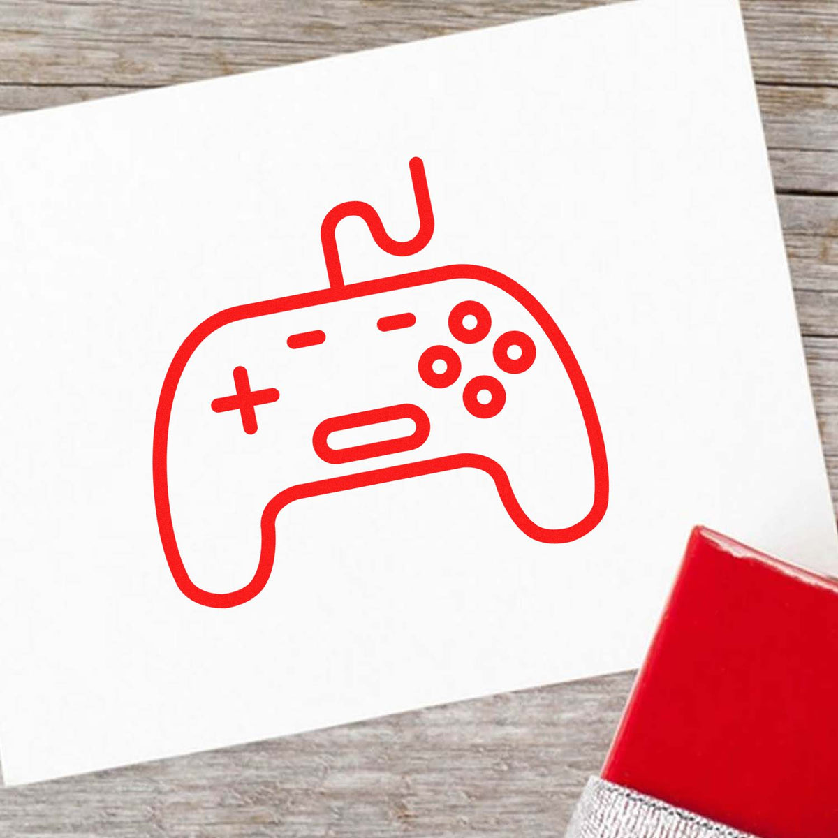 Red game controller-shaped stamp and seal on white card, with partial red stamp block on rustic wood background.