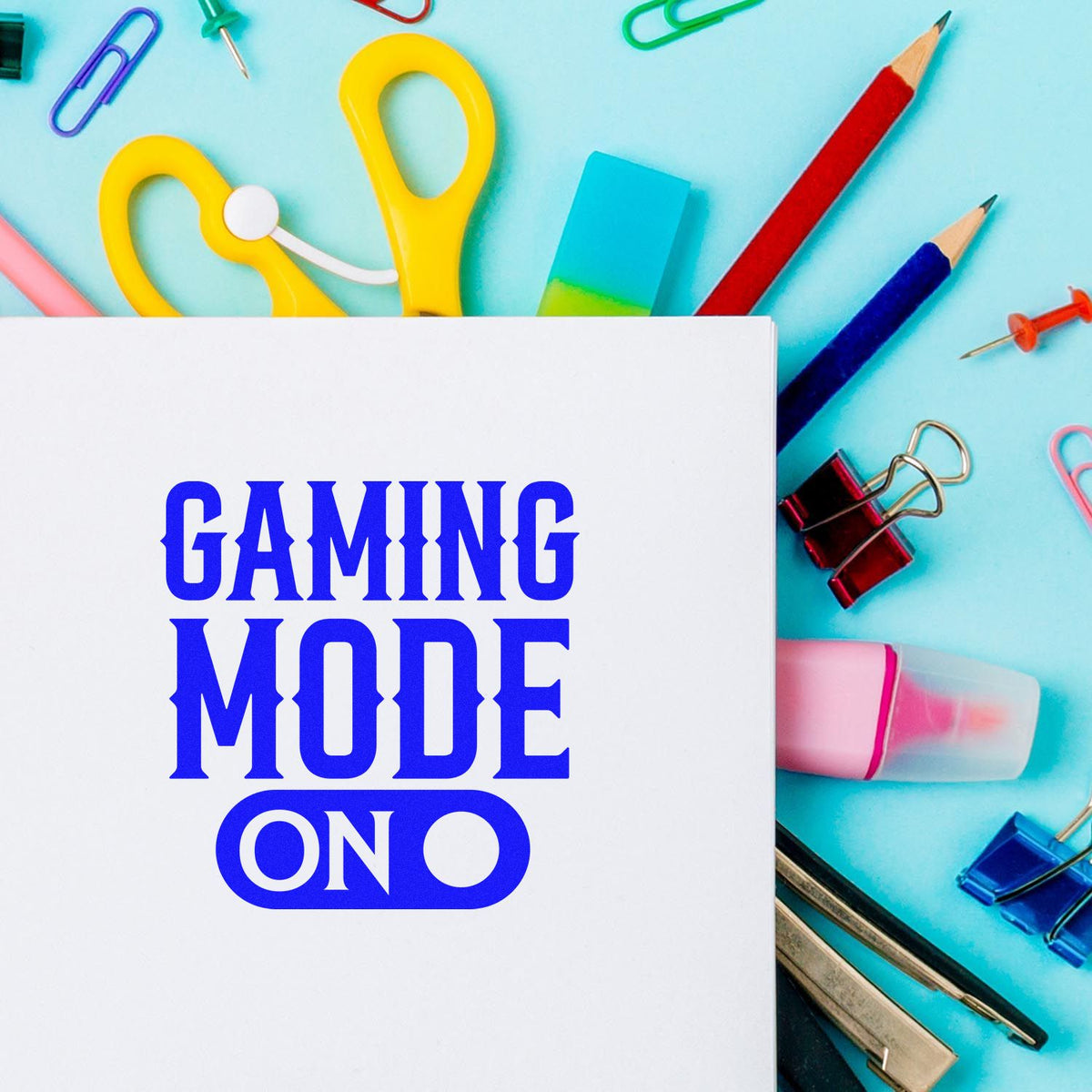 Blue GAMING MODE ON stamp/seal on white card, with colorful stationery, yellow scissors, pencils, and binder clips on turquoise background.