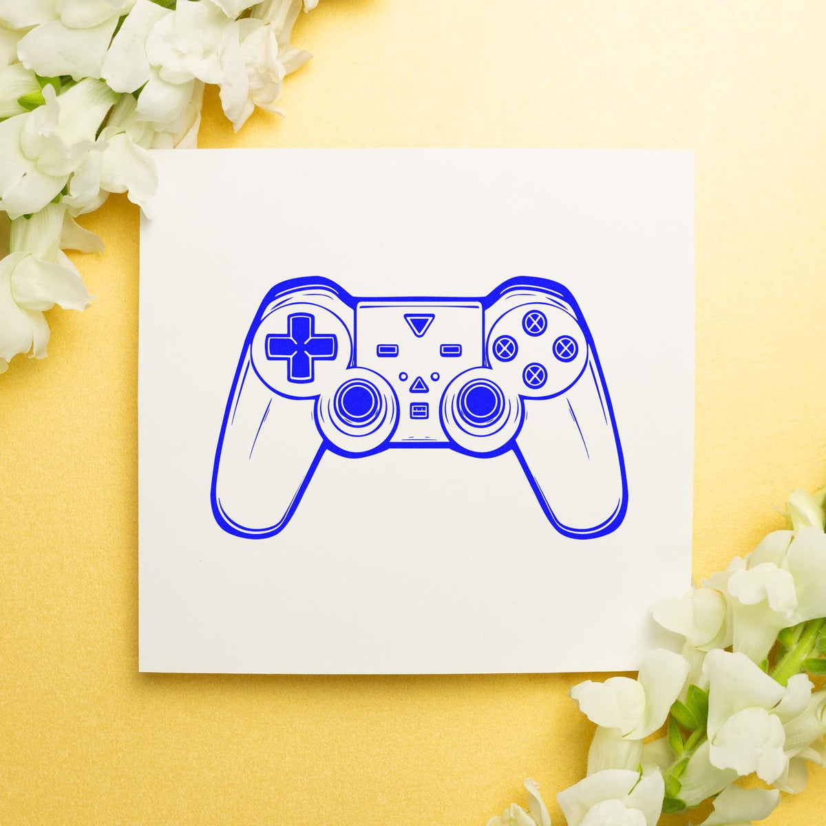 Blue game controller stamp-and-seal design on white card, framed by white flowers over a soft yellow background.