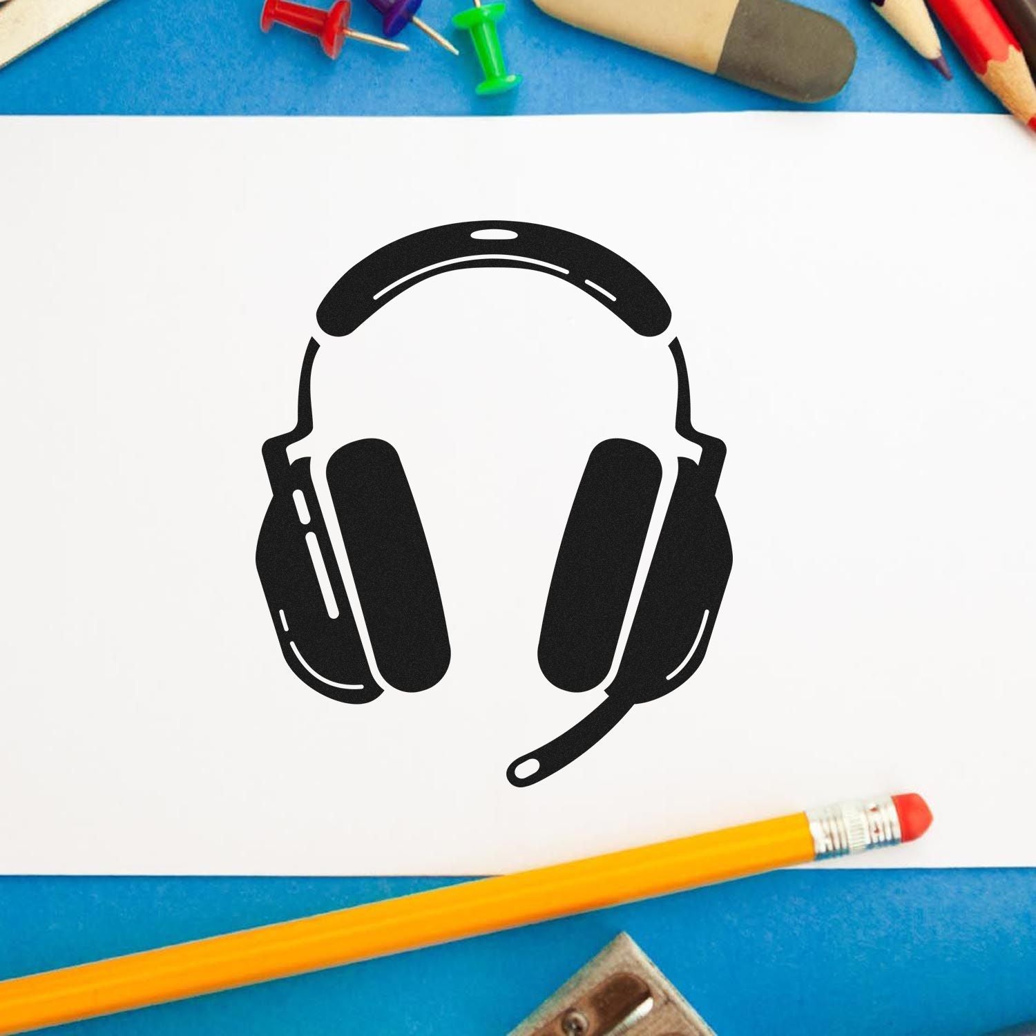 Black headset stamp/seal on white paper, surrounded by colorful pencils, pushpins, and blue background.