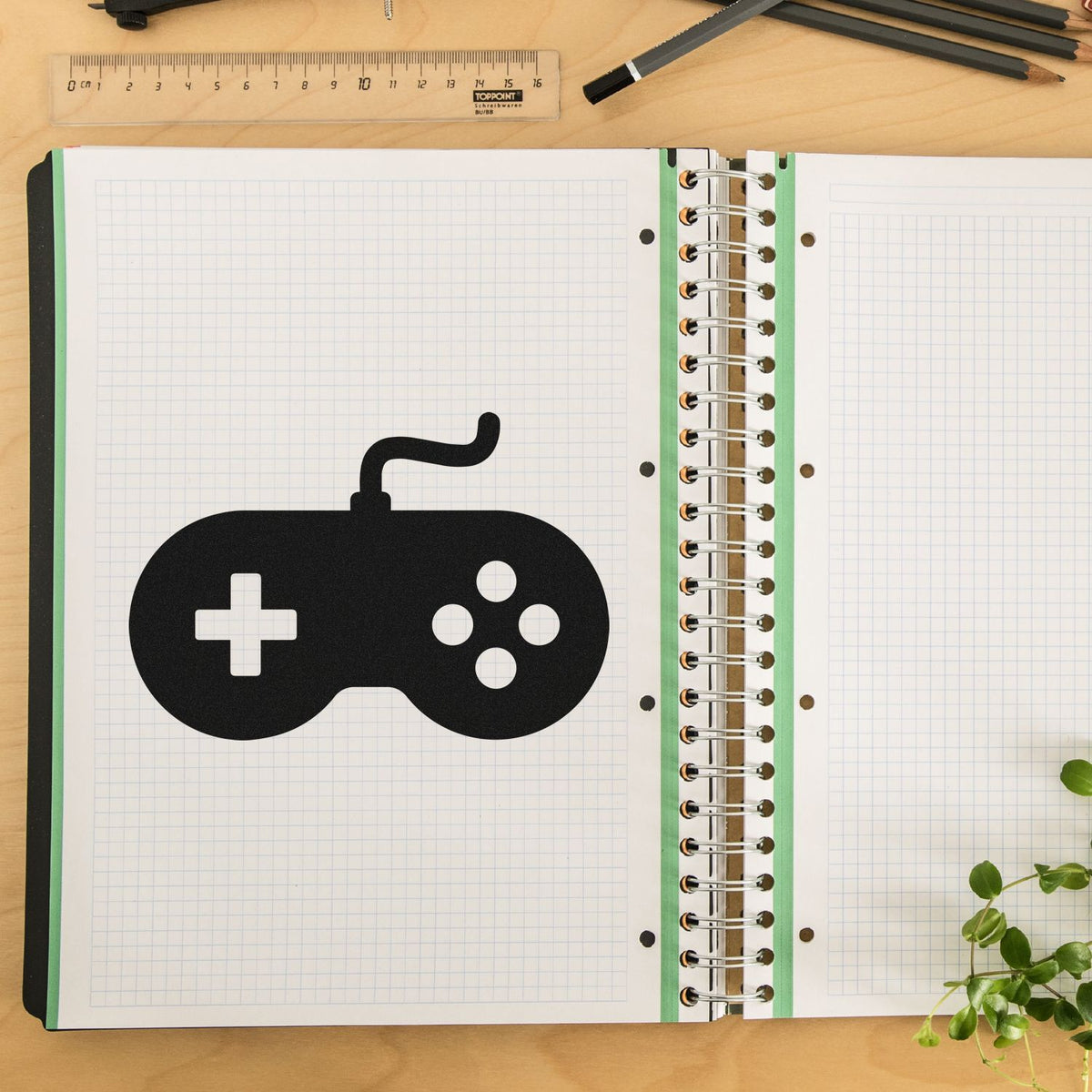 Black controller-shaped stamp and seal on graph-paper notebook, with spiral binding, ruler, pencils, and green plant.