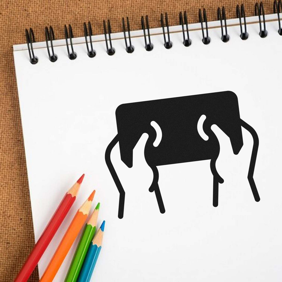 Black stamp and seal product graphic on white spiral notebook, with colorful pencils on a tan background.