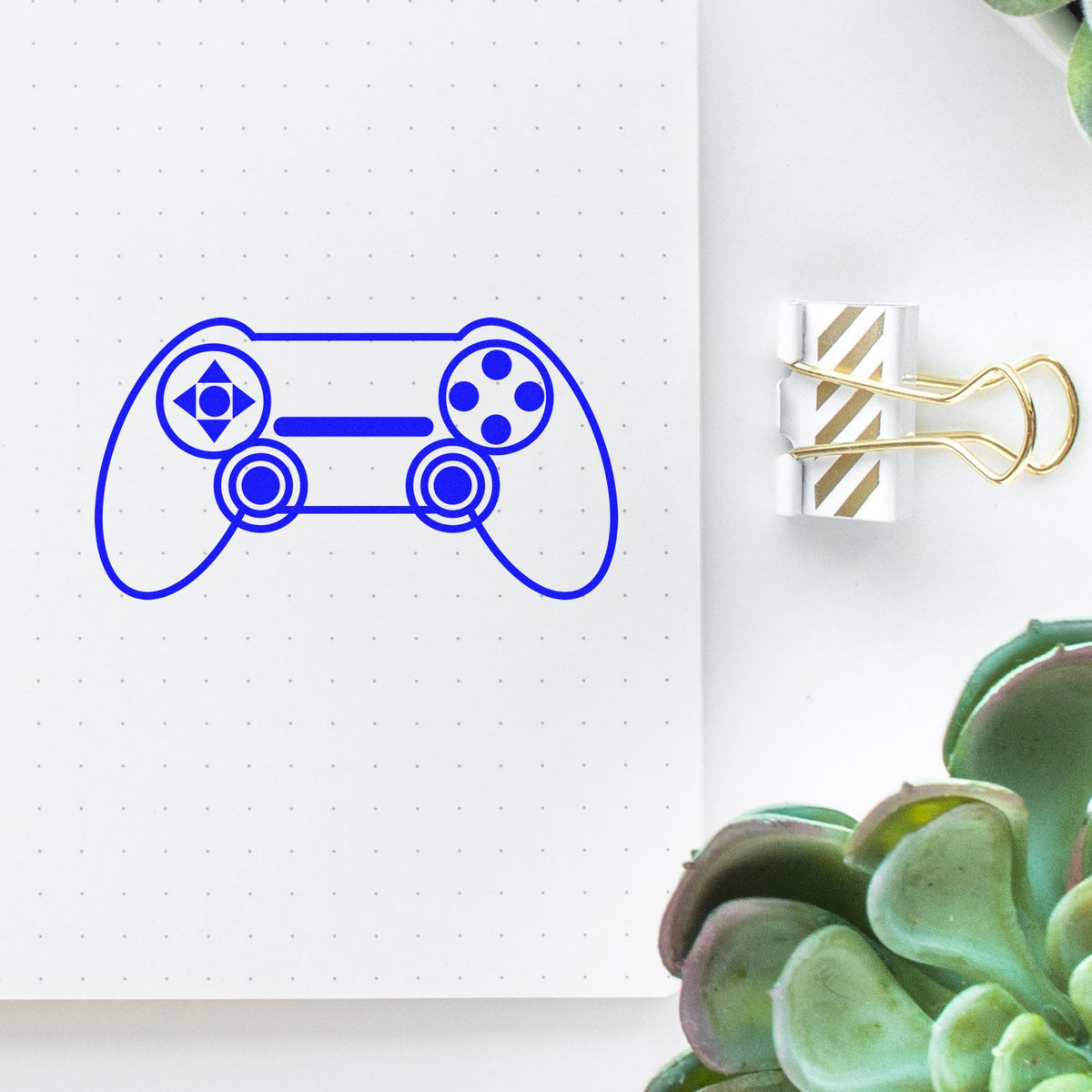 Blue custom stamp seal shaped like a game controller on dotted white paper, with a gold-striped binder clip and green succulent.