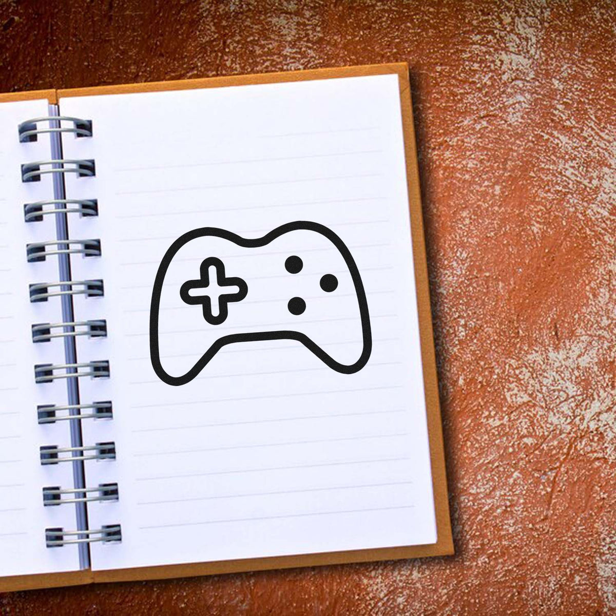 Black game controller stamp/seal on a white spiral notebook, with lined pages and brown textured background.