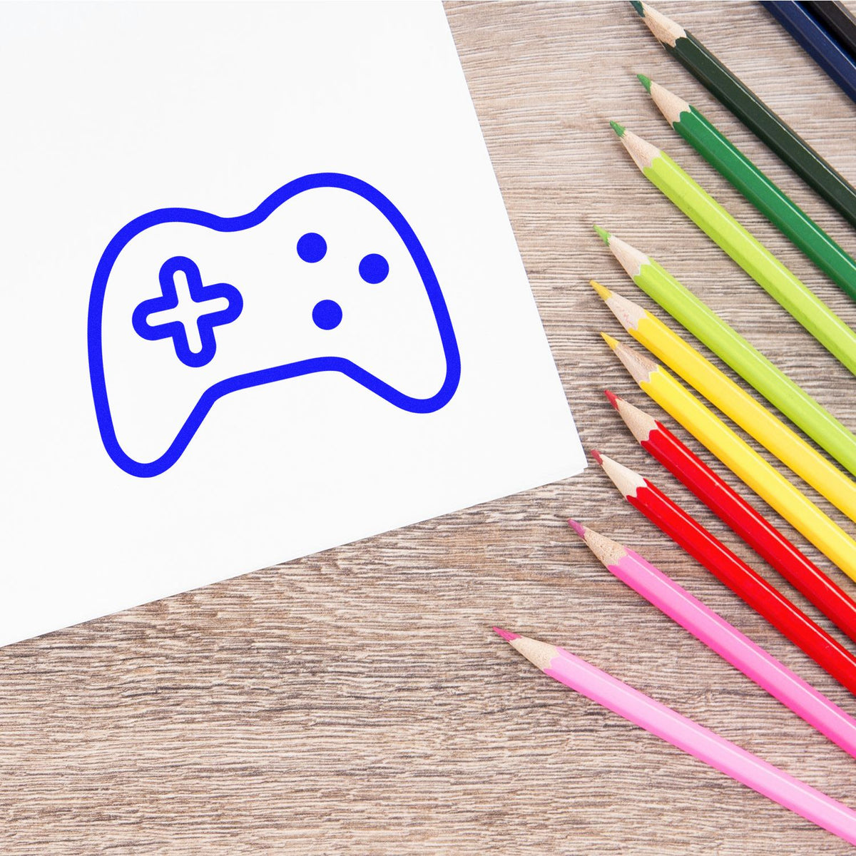 Blue game controller stamp and seal design on white paper, with colorful pencils on a wood desk.