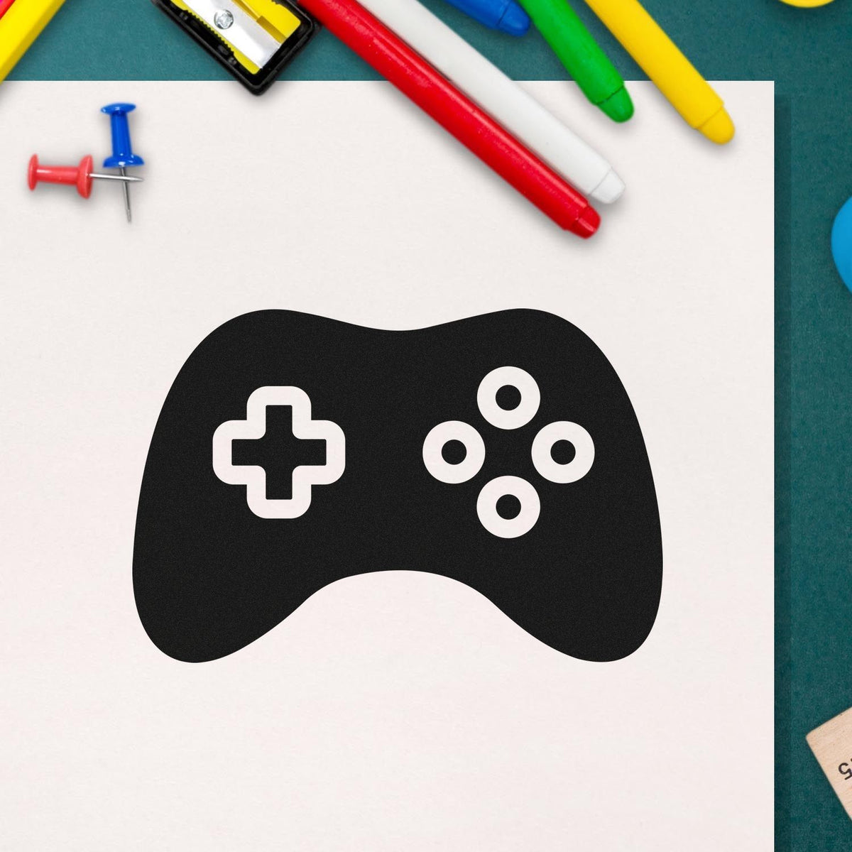 Black game controller-style stamp and seal with white cross and buttons, surrounded by colorful pens and push pins.