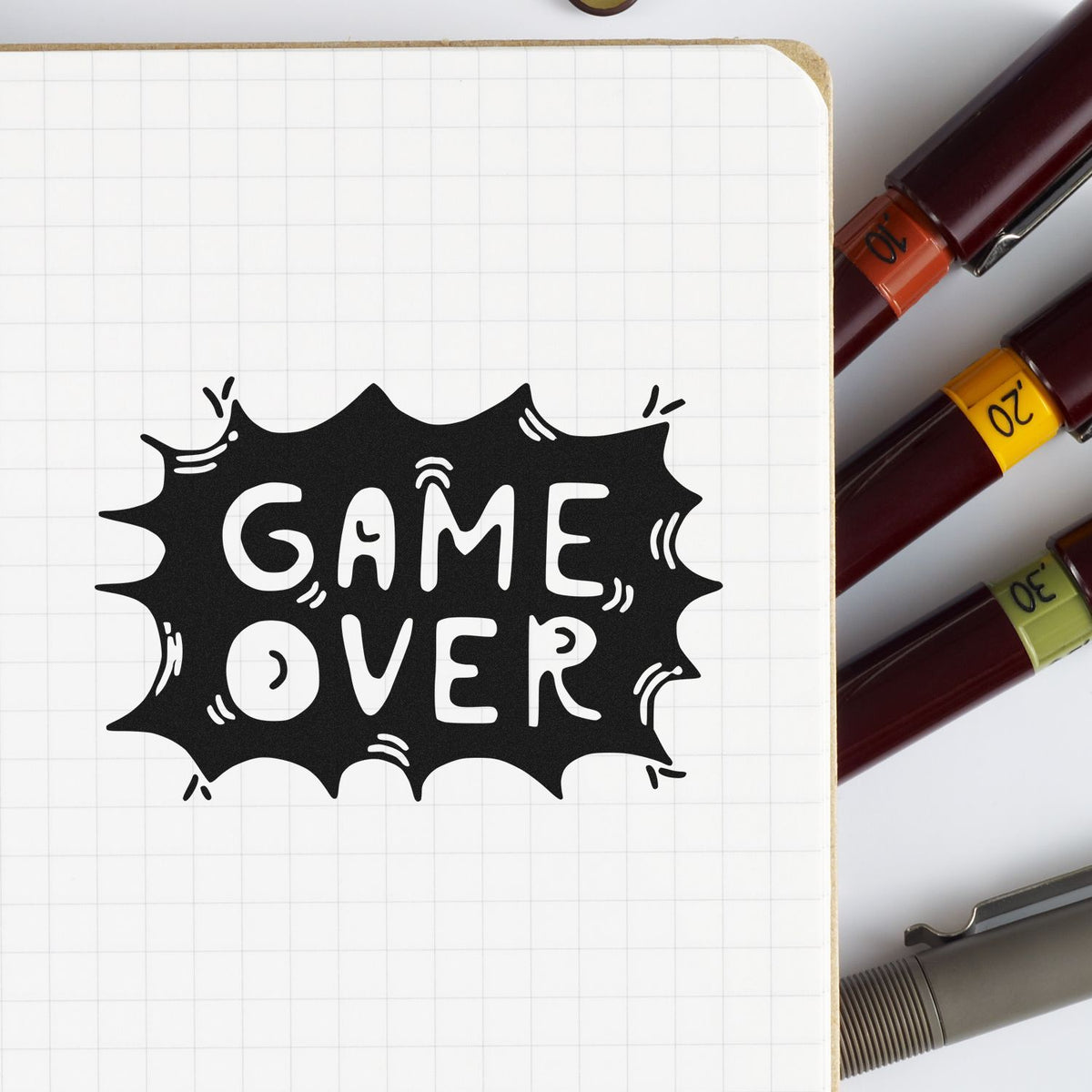Black GAME OVER stamp on a grid notebook, with burgundy seal markers capped orange, yellow, and green.
