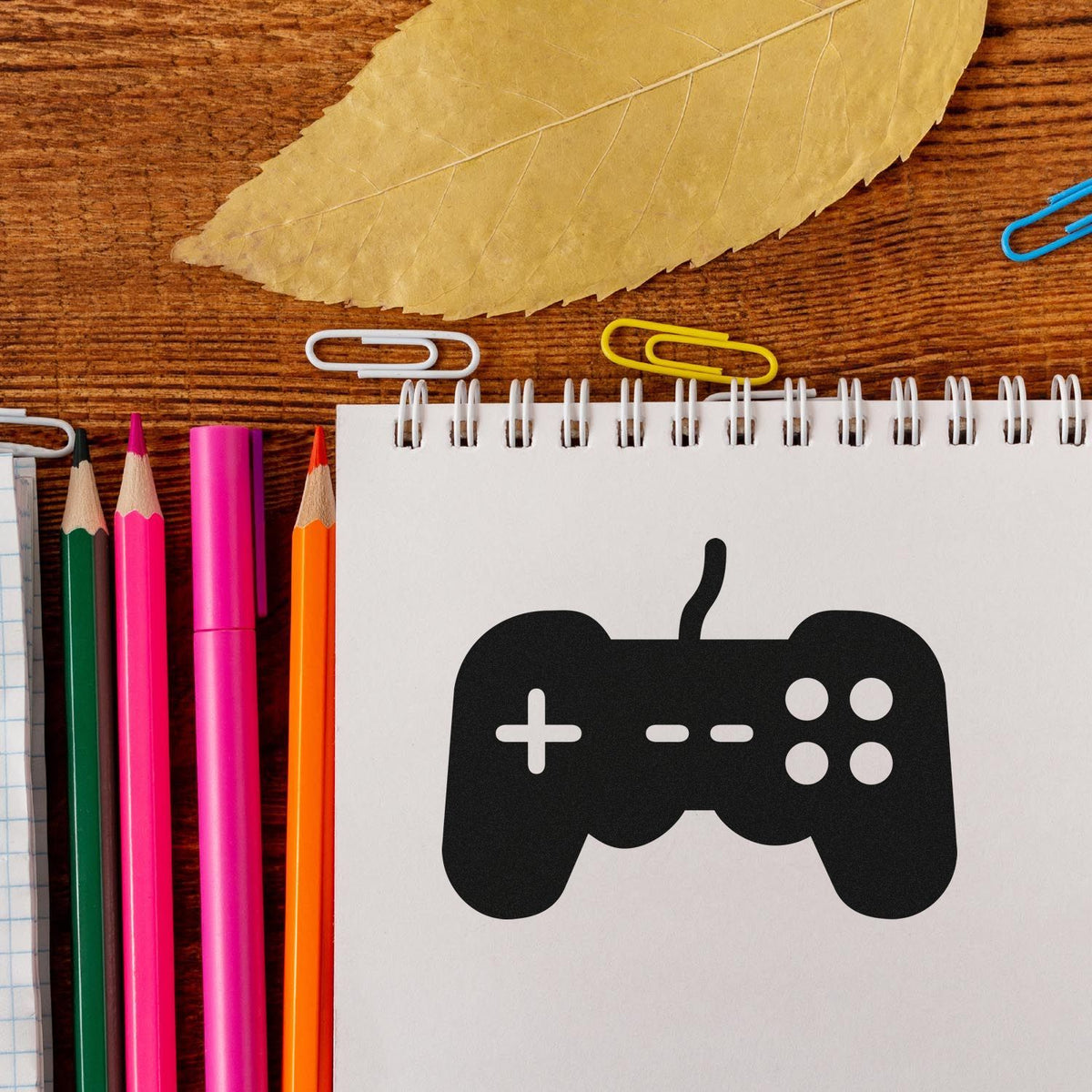 Black game controller stamp and seal on white spiral notebook, with pink, orange and green pencils, paper clips, and a leaf.