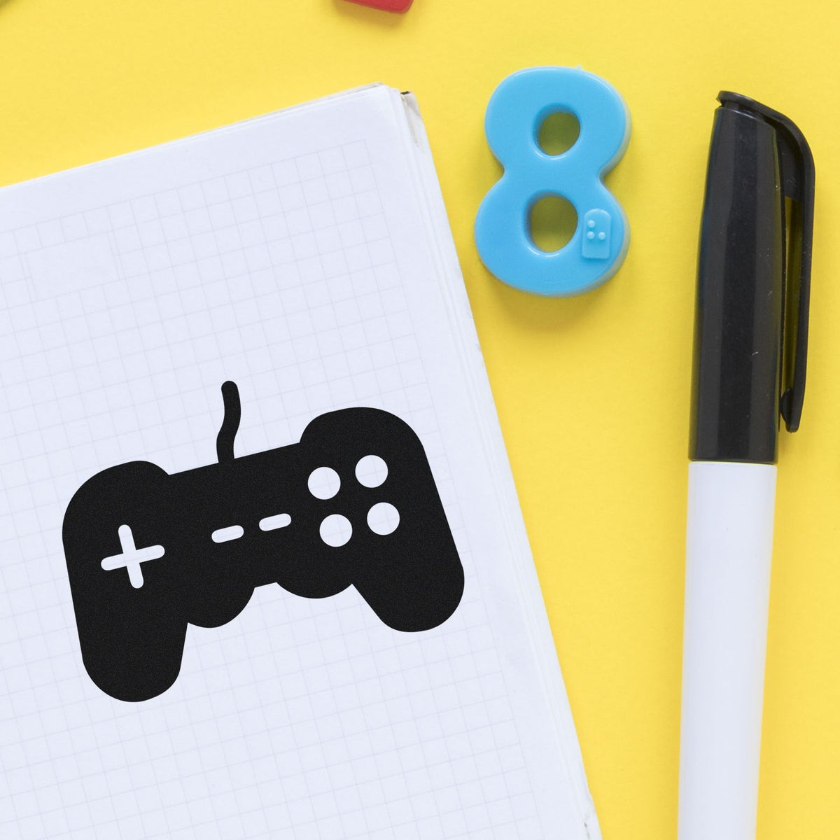 Blue number 8 stamp, black game controller seal on graph notebook, with black marker on bright yellow background.