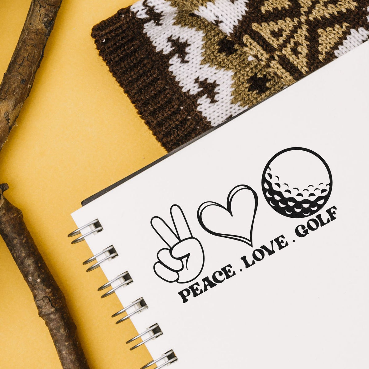 White spiral notebook with black peace-love-golf stamp seal, beside brown knit swatch and wood sticks on a yellow background.