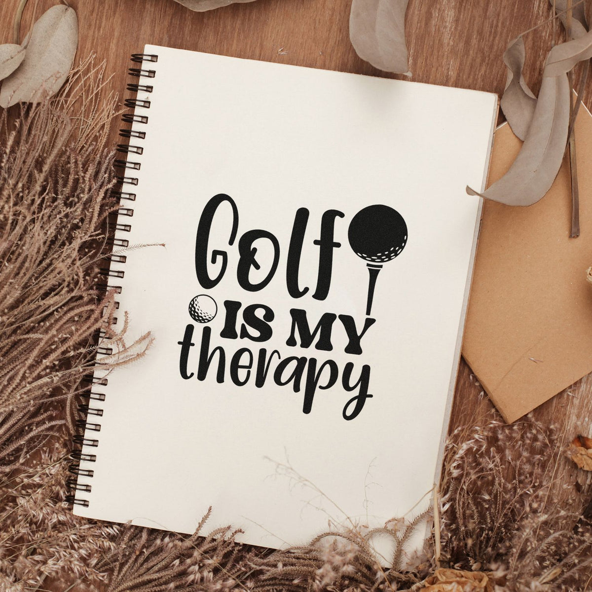 Black golf-themed stamp/seal on a cream spiral notebook, with golf ball and tee graphic, on warm wood with dried beige decor.