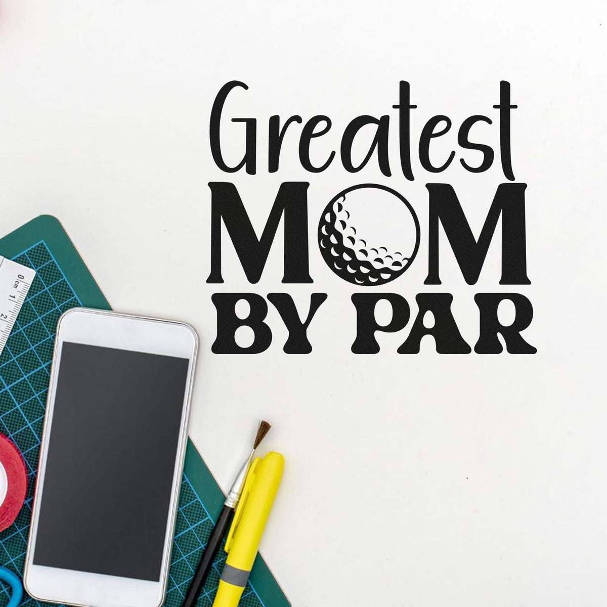 Black "Greatest Mom by Par" stamp and seal design with golf ball O, on white craft desk with cutting mat, phone, and tools.