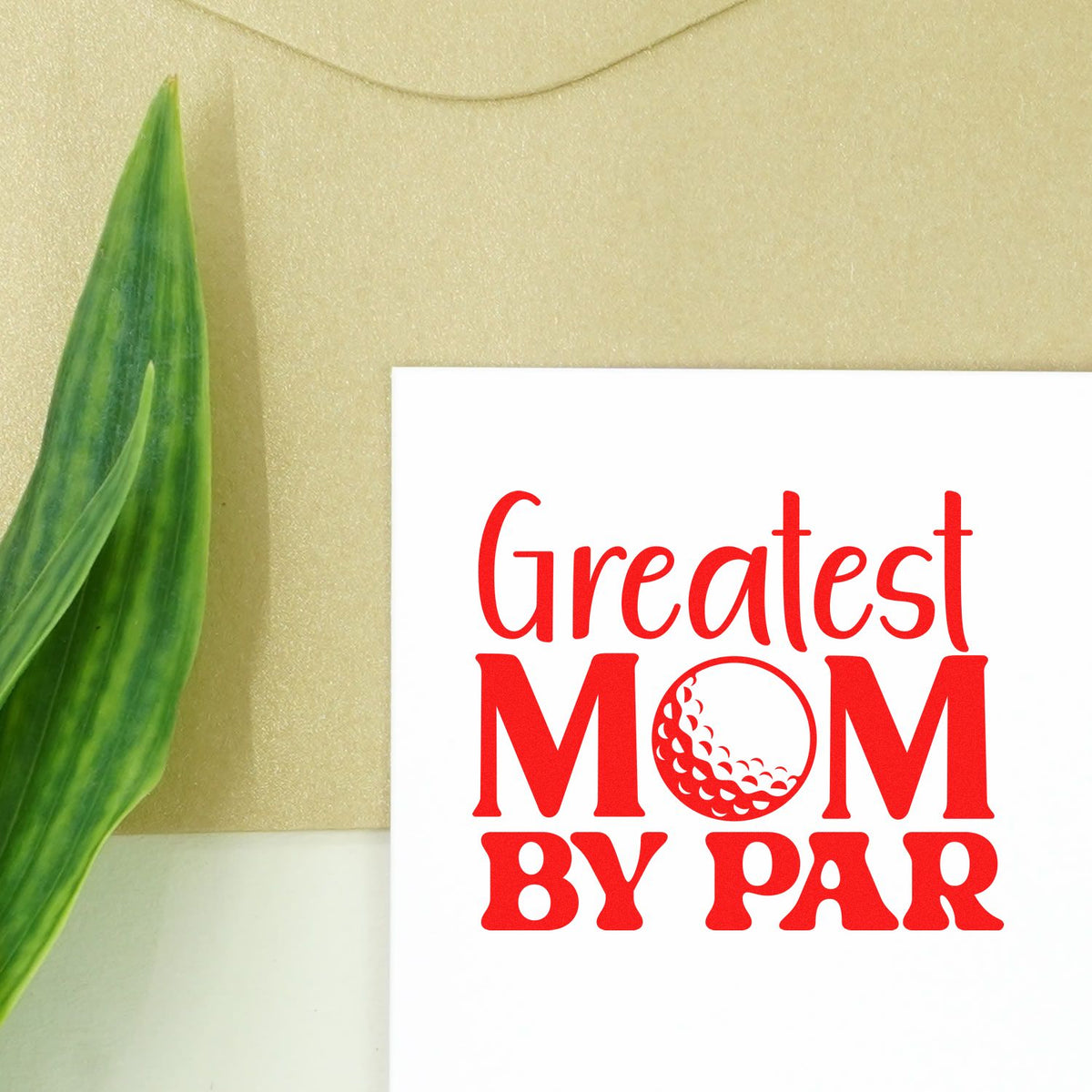 Red Greatest Mom by Par stamp/seal design on white card beside a green leaf, beige background