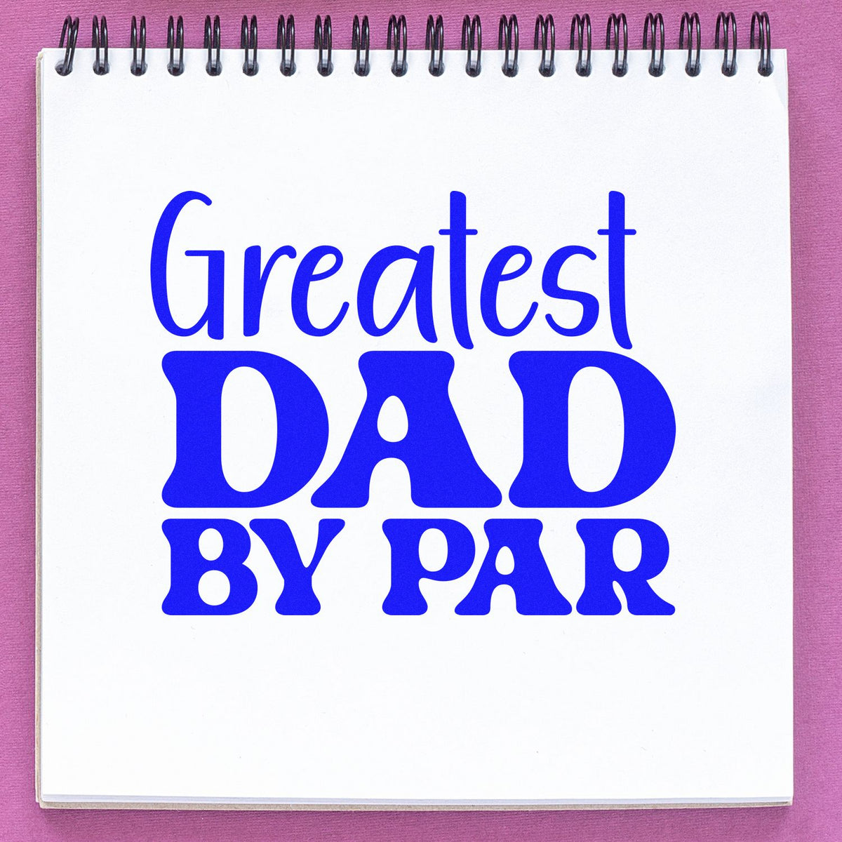 Blue Greatest DAD BY FAR rubber stamp seal on a white spiral notepad, set against a pink background.