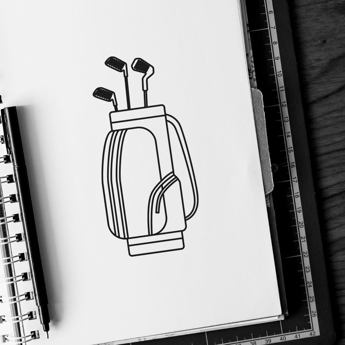 Black-and-white golf bag stamp and seal with clubs on sketchbook, beside pen and ruler