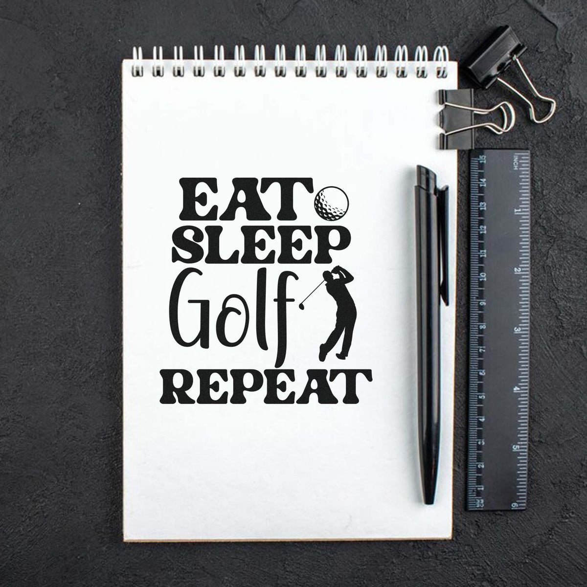 Black golf stamp and seal design on white spiral notebook, with ruler, pen, and binder clips on dark background.