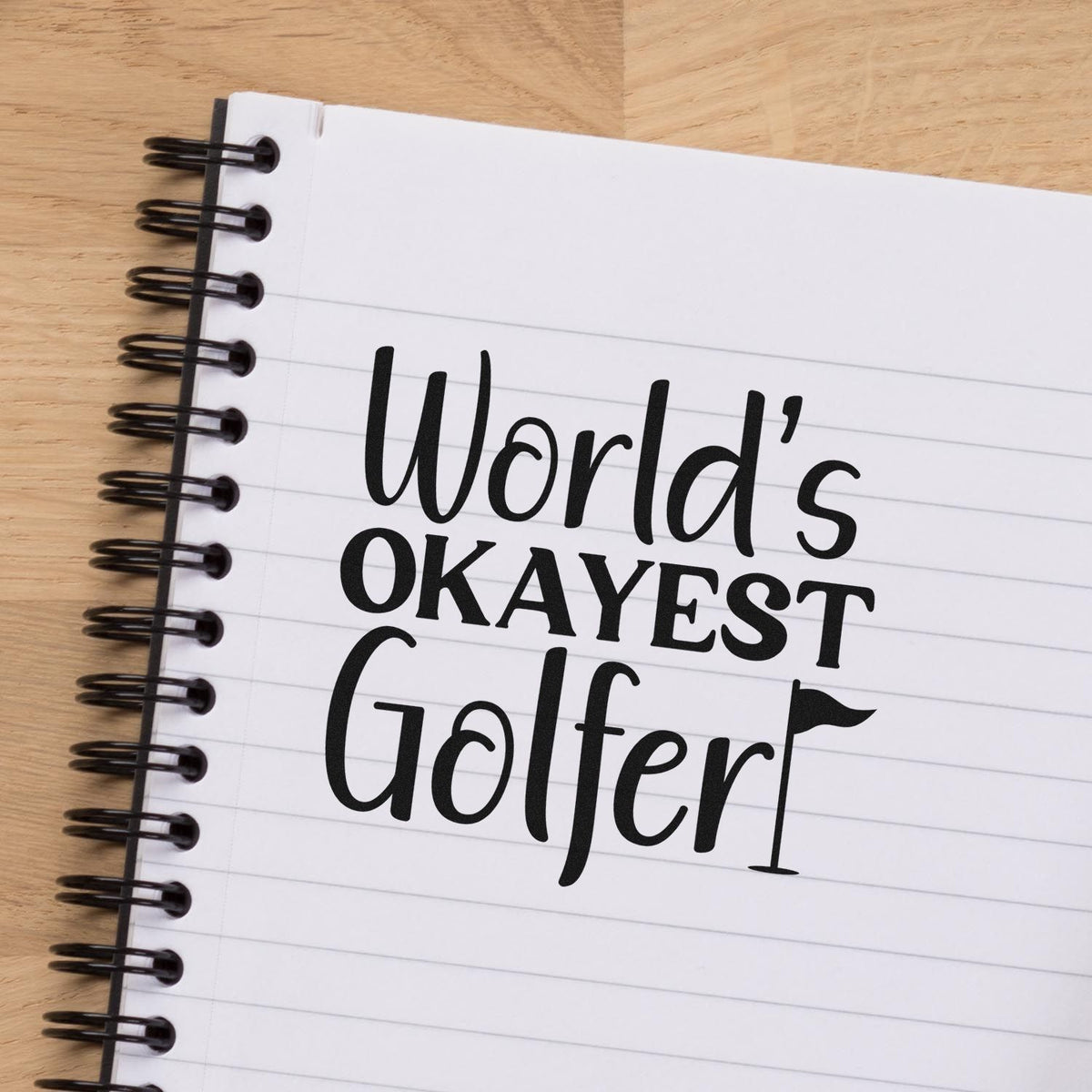 Custom black stamp/seal reading Worlds Okayest Golfer on a white spiral notebook with lined pages and wood background.