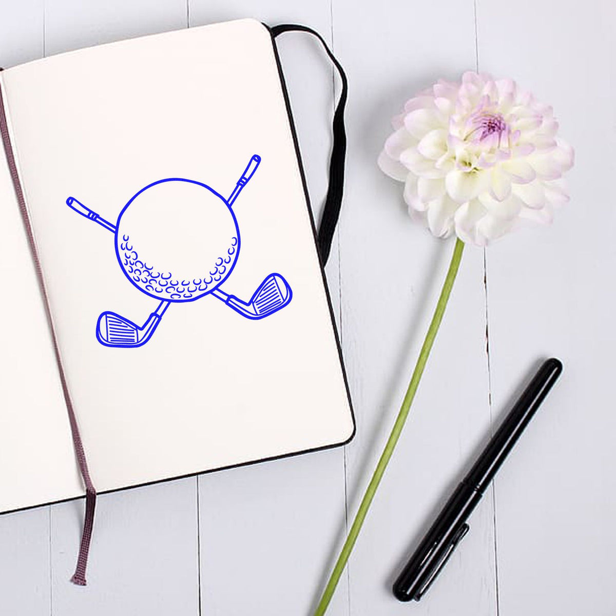 White notebook featuring a blue golf ball and clubs stamp design, beside a black pen and white flower on pale wood.