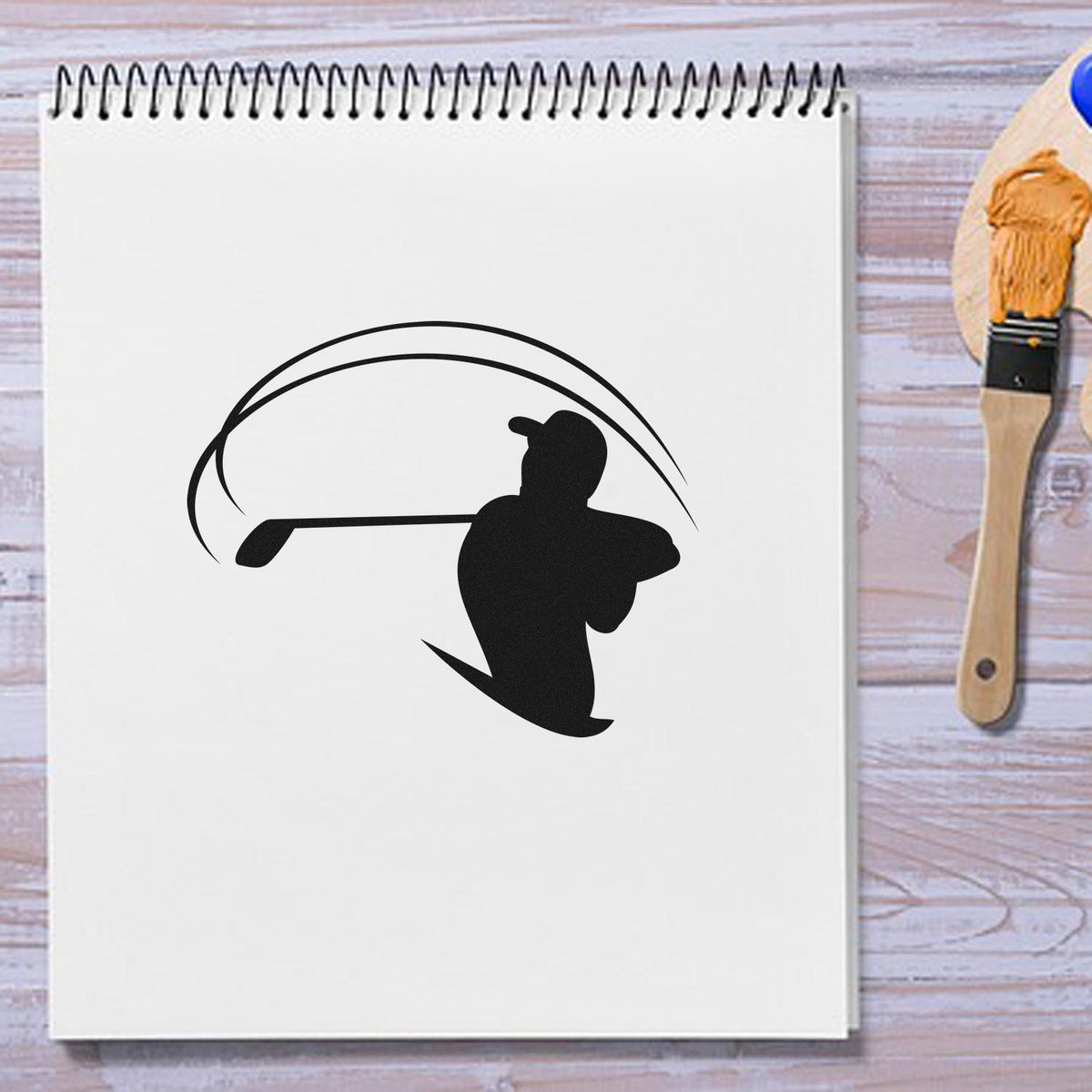 Black golfer silhouette stamp and seal imprint on a white spiral notebook, with a wooden paintbrush beside it.