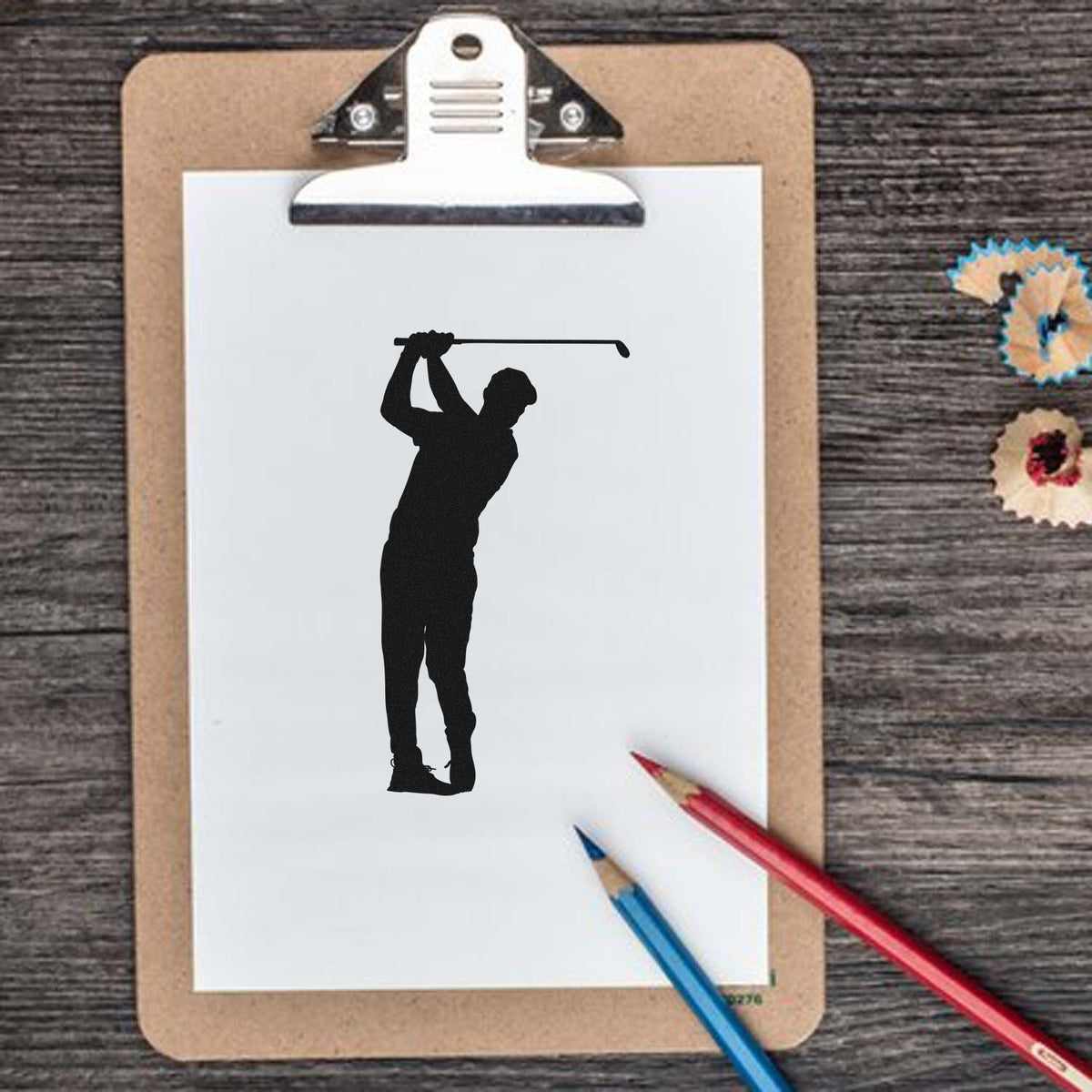 Black golfer silhouette stamp on white paper clipped to tan clipboard, with red and blue pencils on rustic wood and seal samples