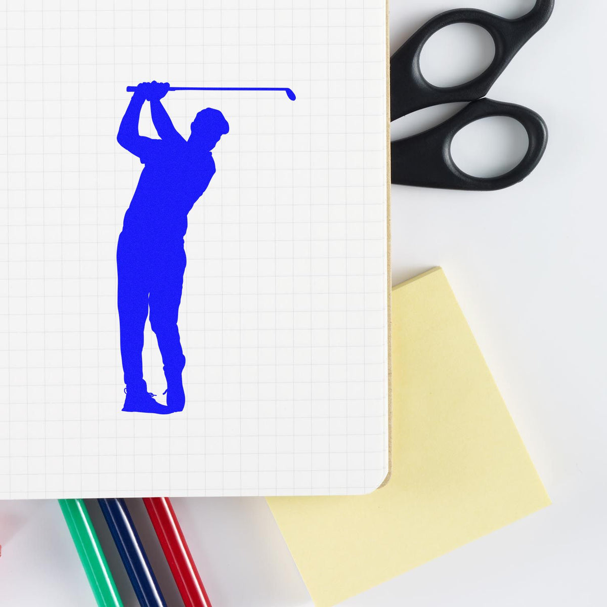 Blue golfer stamp/seal on grid notebook with black scissors, yellow sticky note, and colored pens.