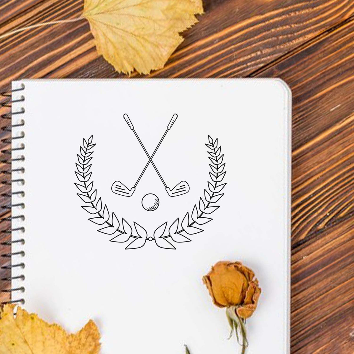 White spiral notebook with black golf club laurel seal stamp on a wooden table, accented by dried yellow leaves and flower.