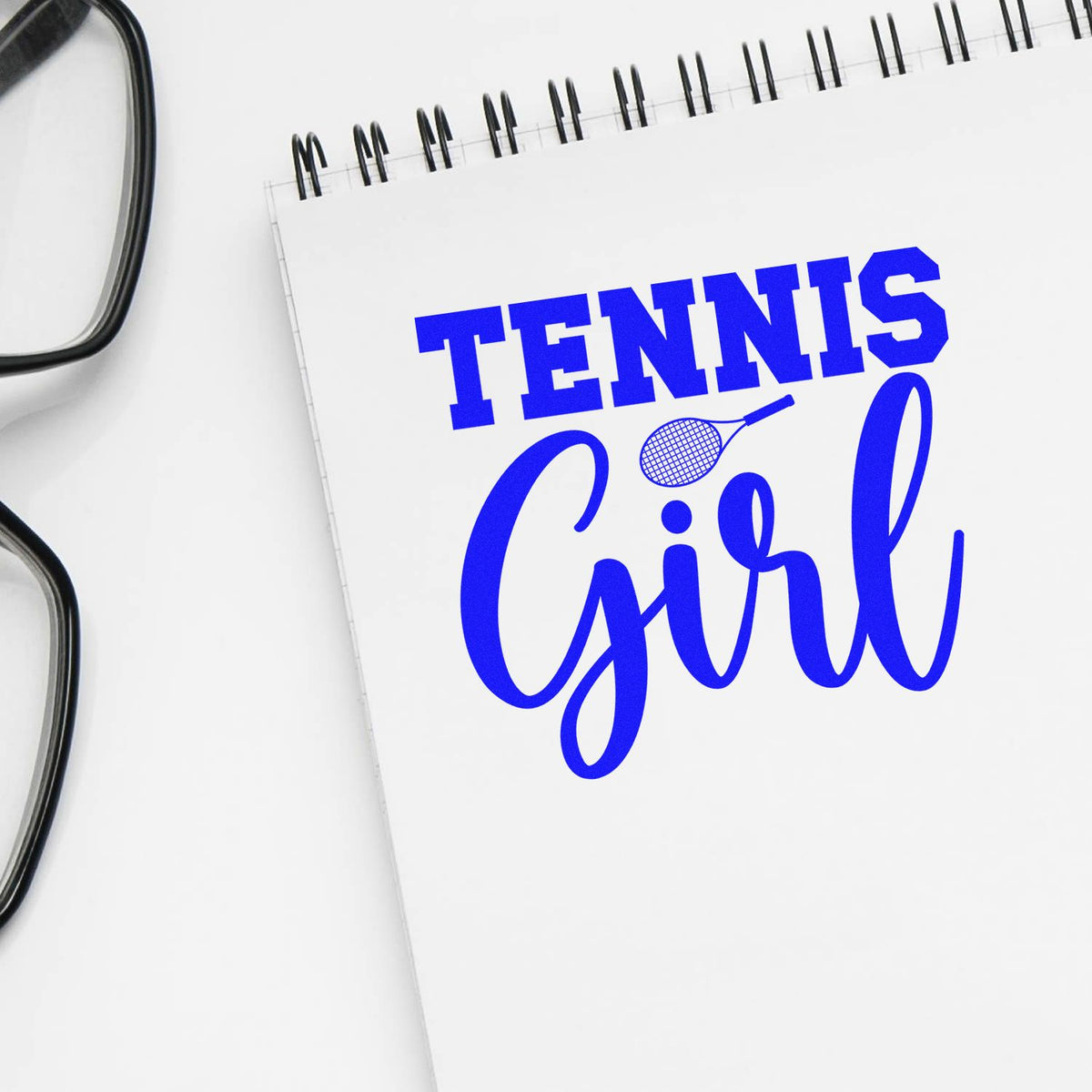 Blue custom stamp and seal reading Tennis Girl with tennis racket icon on a white spiral notebook, beside black eyeglasses.
