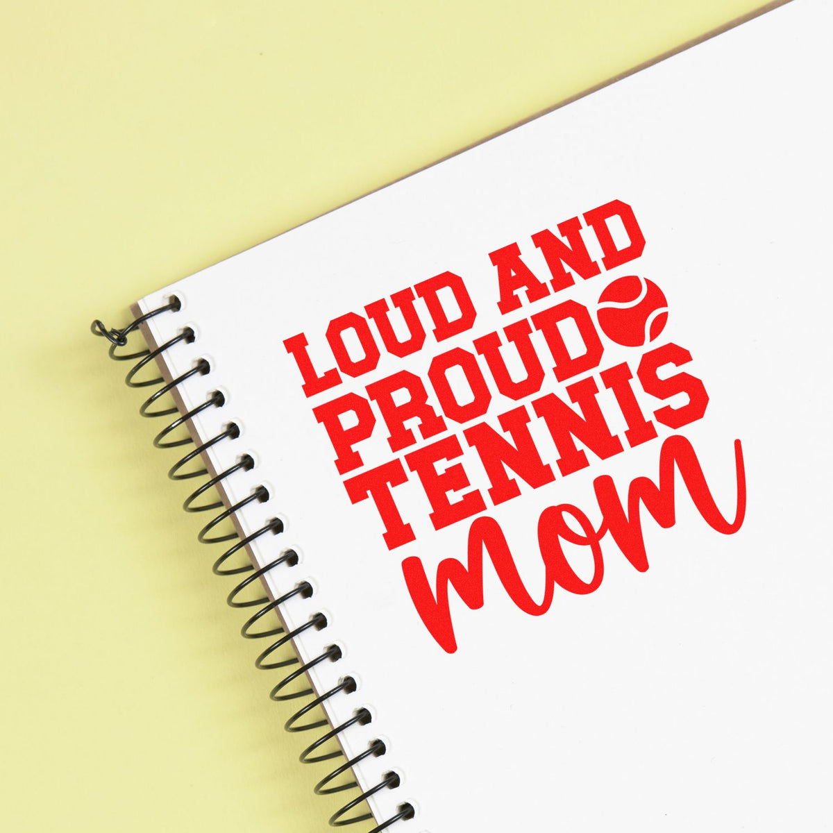 Red Loud and Proud Tennis Mom stamp design on white spiral notebook with black coil, on pale yellow background.