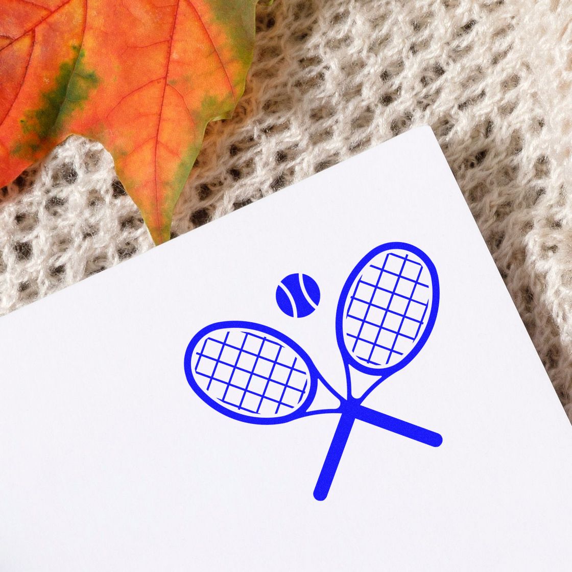 Blue tennis racket and ball stamp seal on white paper, with autumn leaf and beige knit background.
