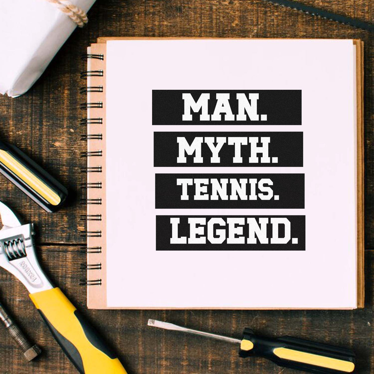 Black-and-white stamp and seal design reading Man. Myth. Tennis. Legend. on a white spiral notebook with tools on a wood table.