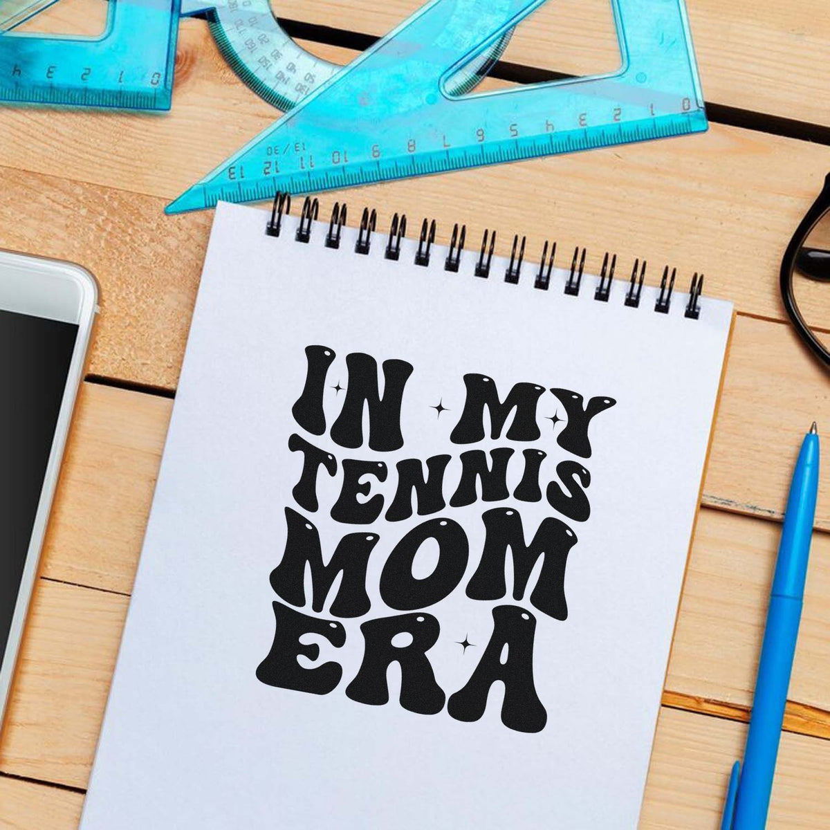 Custom black tennis mom stamp/seal design on white notebook, with blue rulers, pen, and phone on a wood desk.