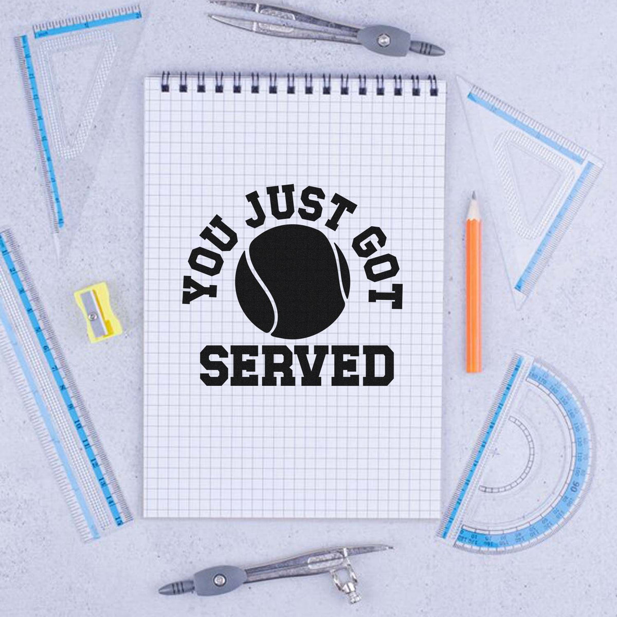 Custom tennis-themed stamp and seal on graph notebook, black YOU JUST GOT SERVED text and ball, with blue rulers and orange pencil.