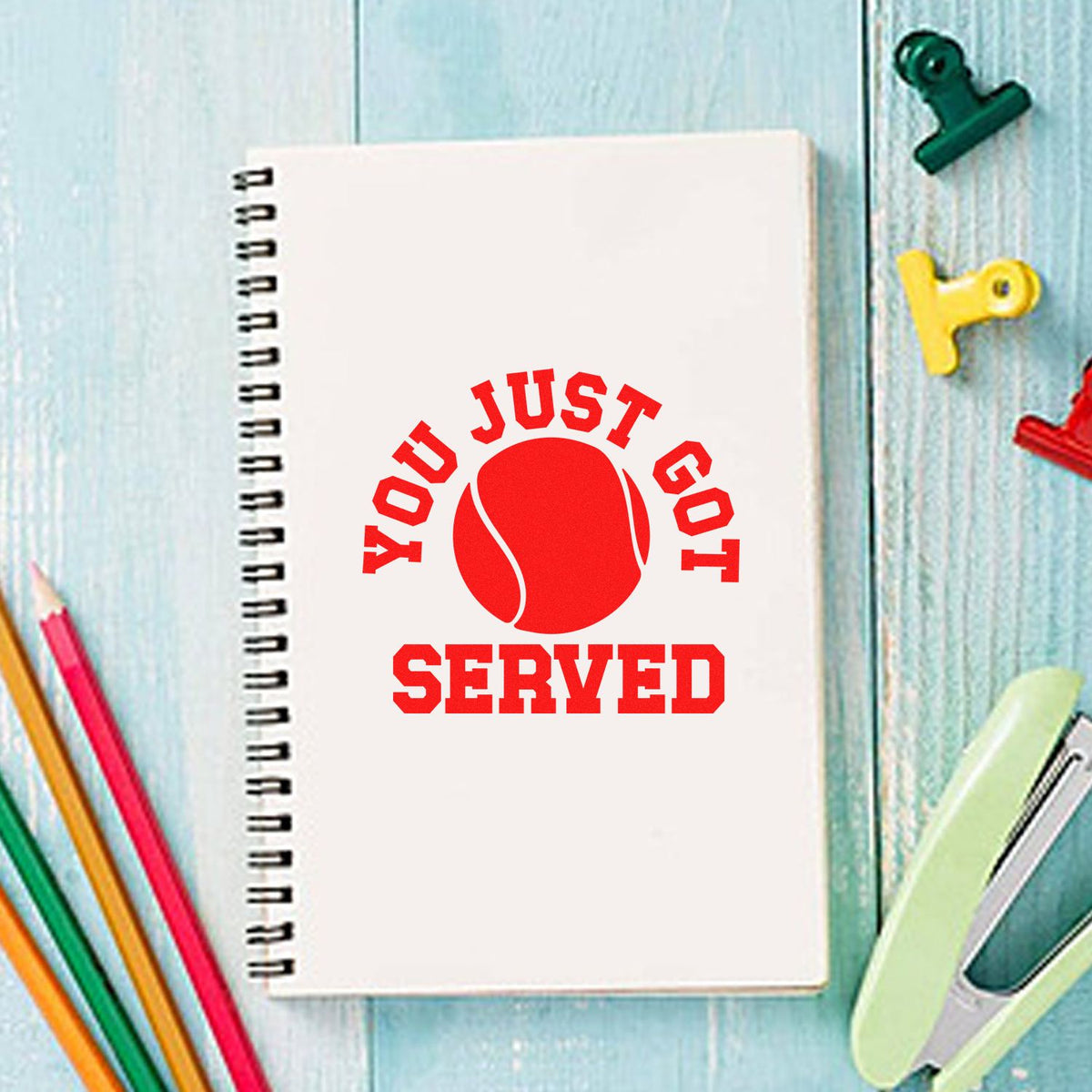 White spiral notebook with red tennis stamp seal reading You Just Got Served, on a light blue desk with colorful stationery.