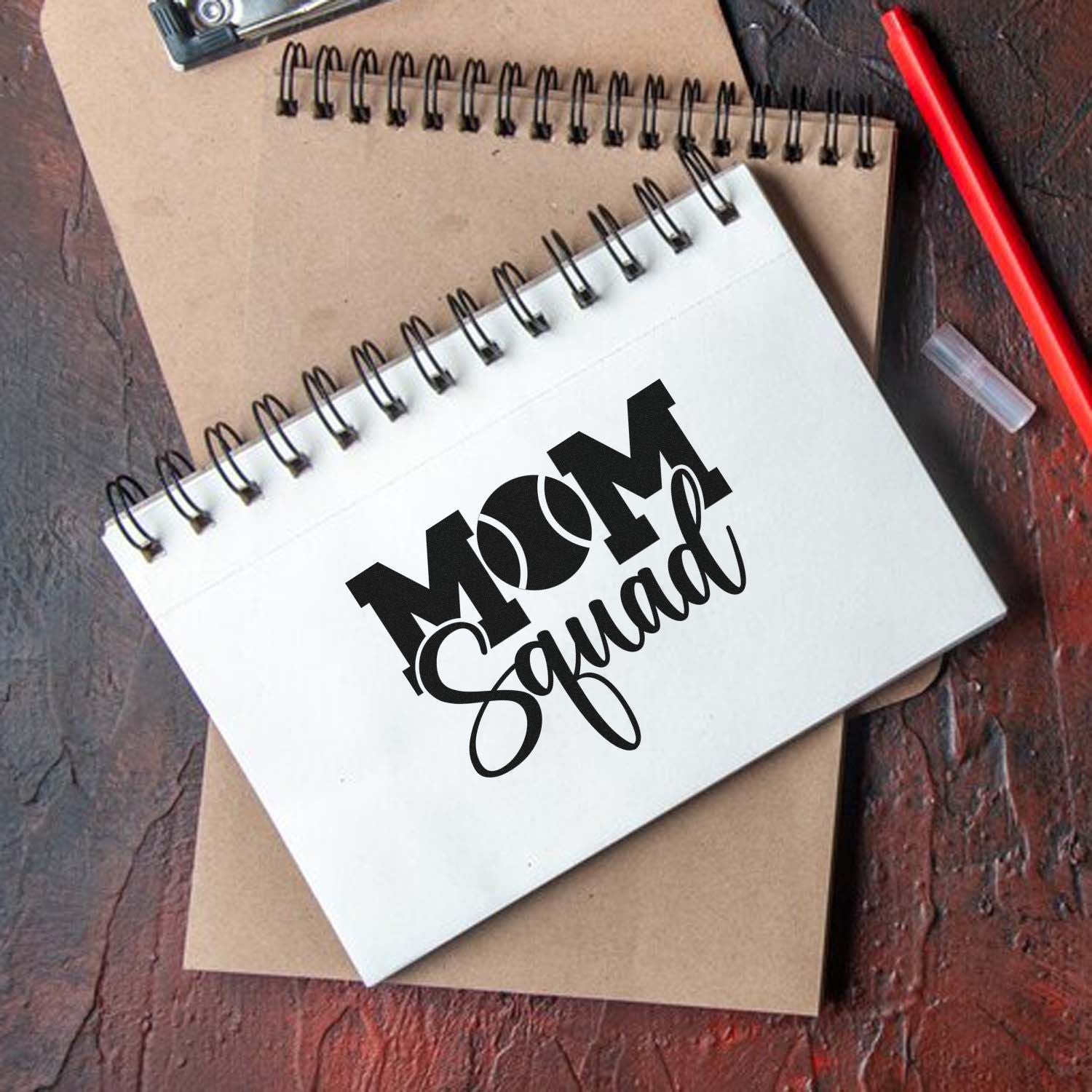 Black MOM Squad custom stamp and seal on white spiral notebook, with tan notebook, red pen, and cap on rustic wood.