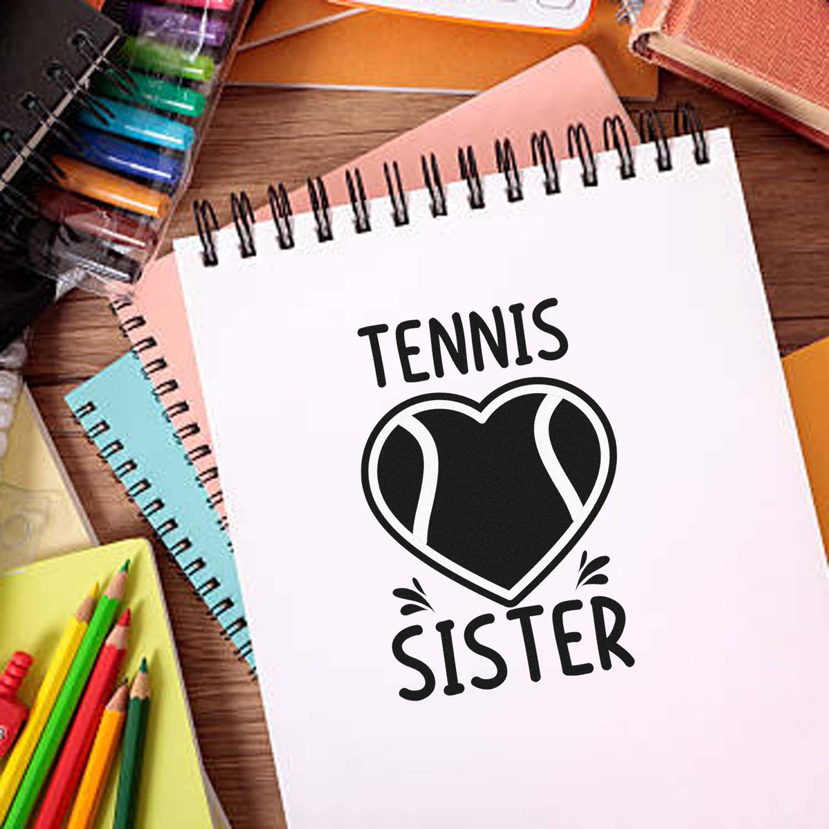 Tennis sister stamp design on a white spiral notebook with a black heart tennis ball graphic, surrounded by colorful notebooks and pencils.