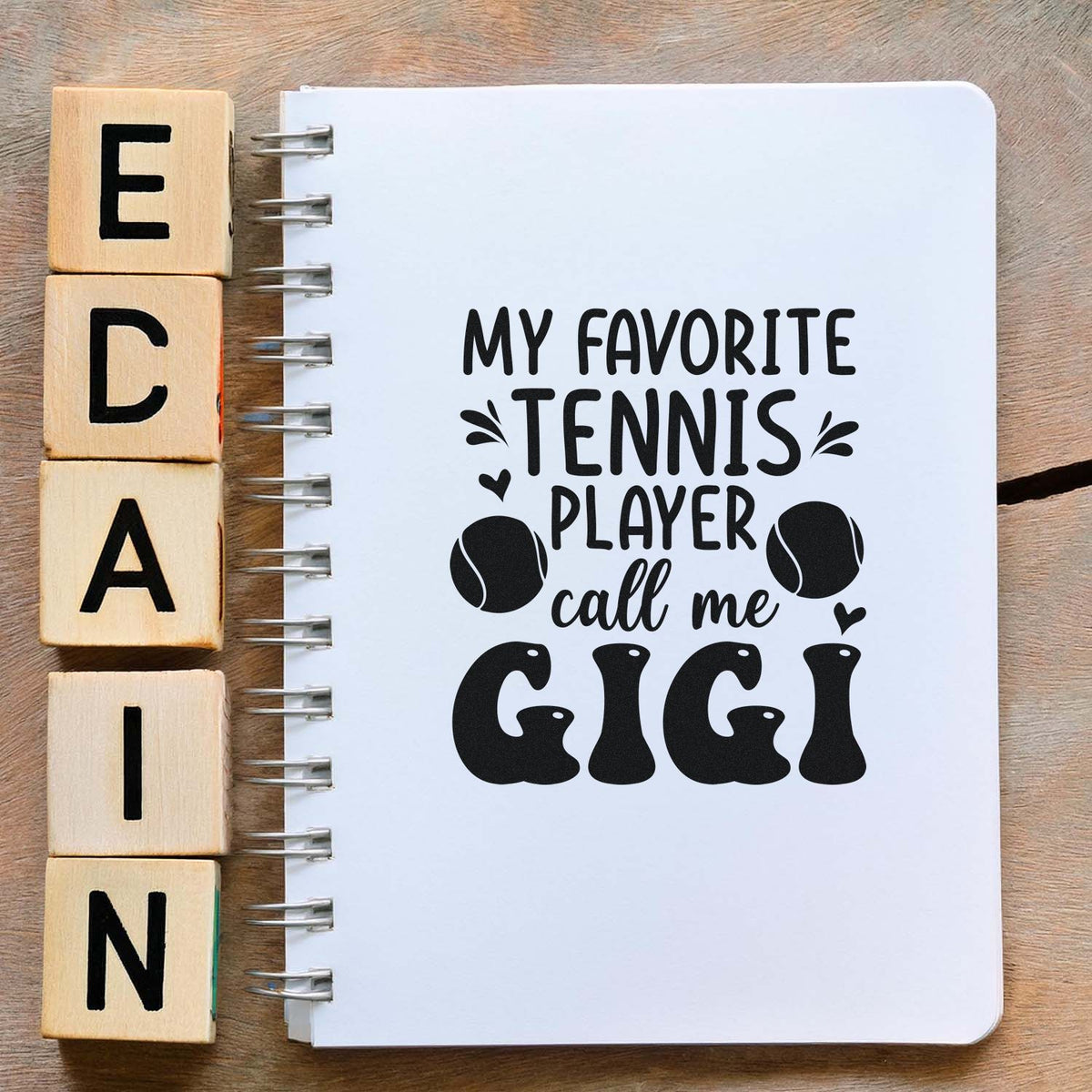 Wooden alphabet stamp blocks beside a white spiral notebook with black tennis-themed call me GIGI design and tennis balls.