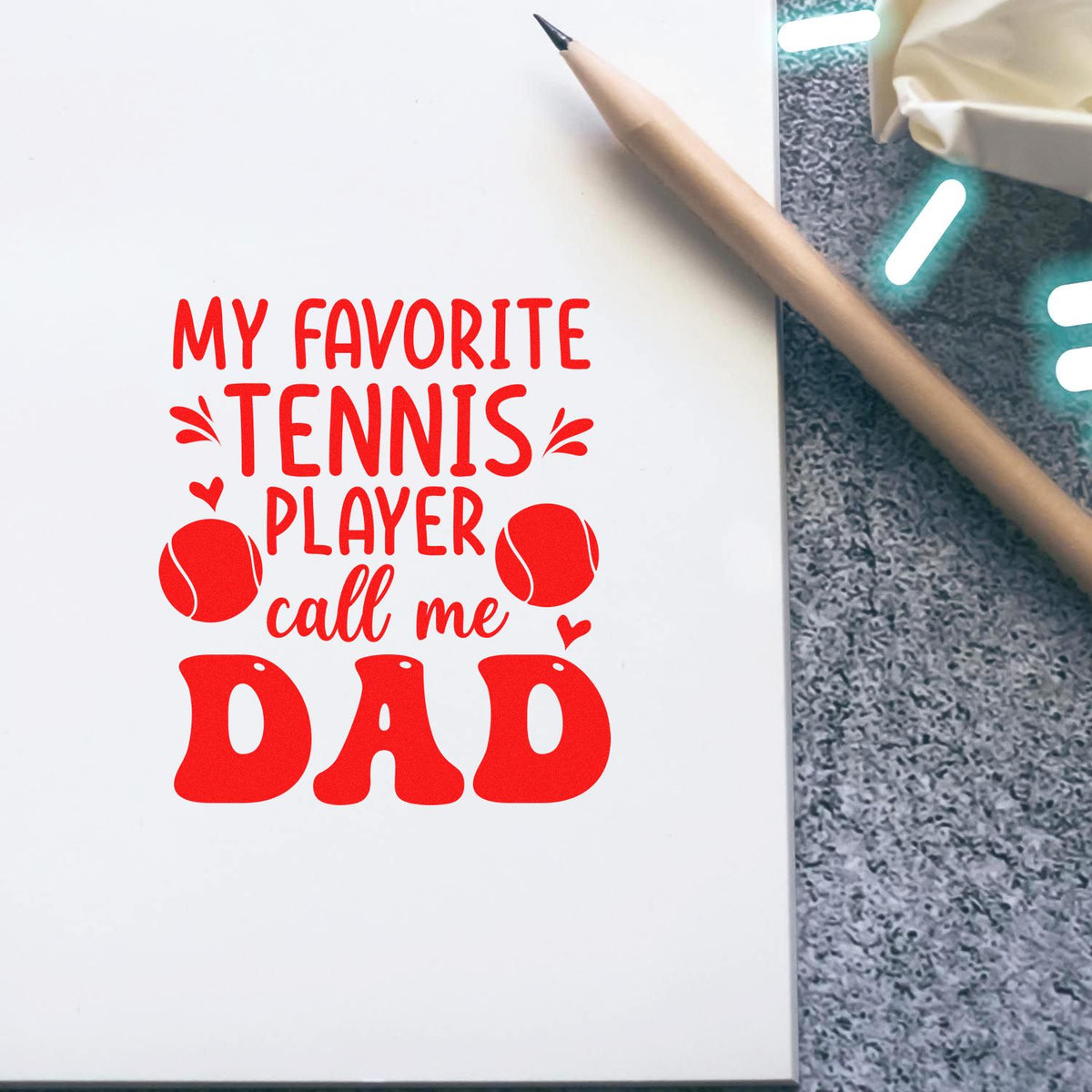 Red tennis stamp design on white page reading My favorite tennis player call me dad, with tennis balls and pencil.