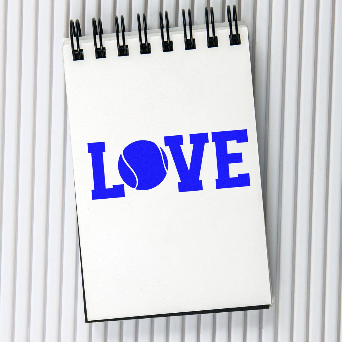 Custom blue LOVE stamp on a spiral notepad, with a tennis ball as the O, against a white ribbed background.
