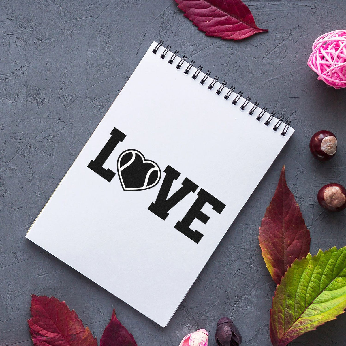 Black LOVE heart stamp seal on a white spiral notebook, with red and green autumn leaves on a gray background.
