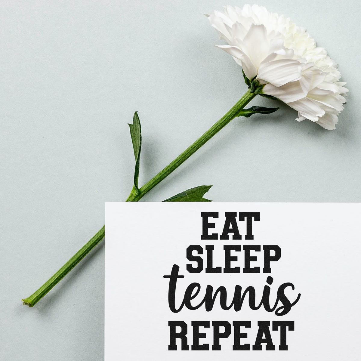 Bold black stamp seal on white card reading Eat Sleep tennis Repeat, beside a white flower with green stem.