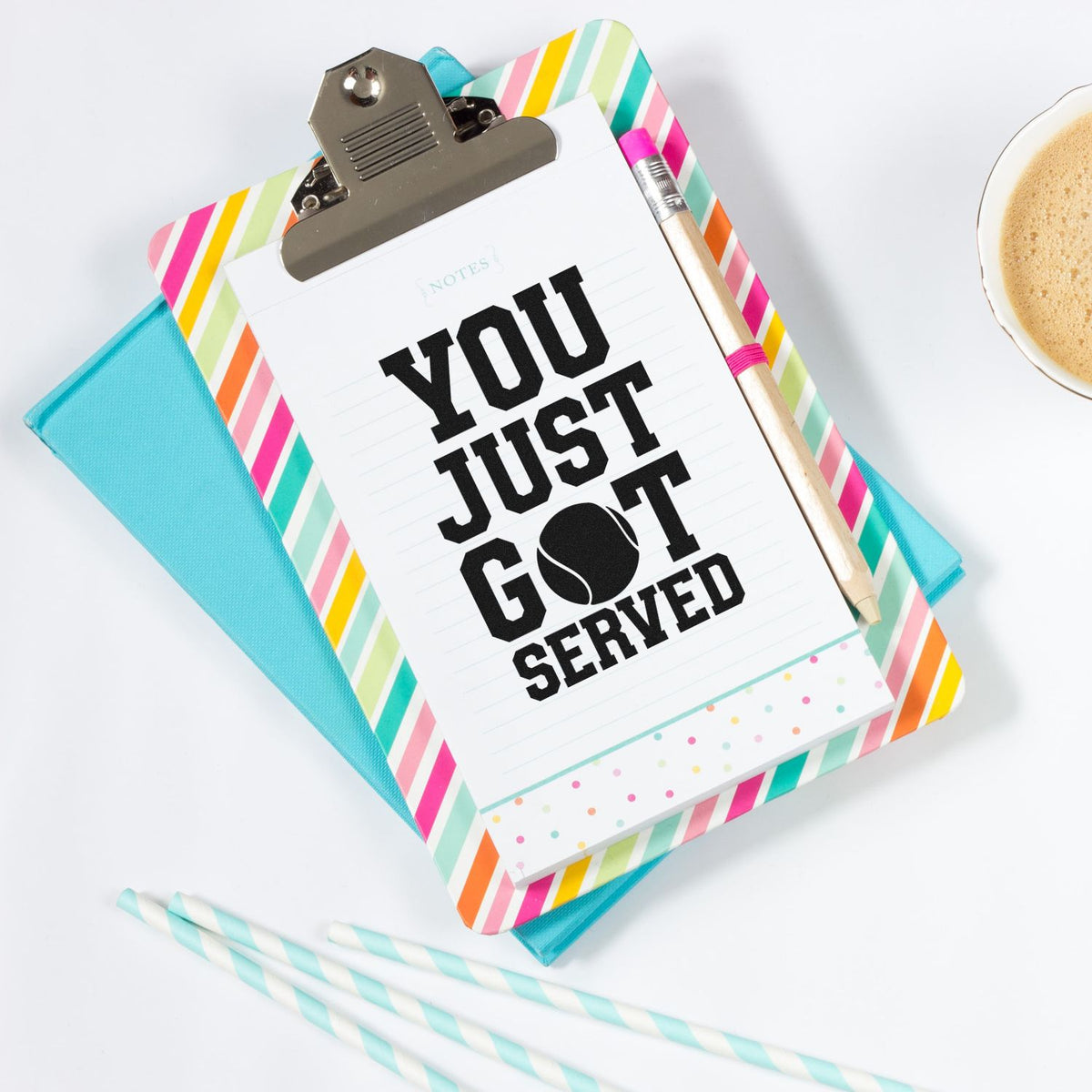 Colorful striped clipboard showcasing a tennis-themed stamp-and-seal reading You Just Got Served, with pencil and turquoise notebook.