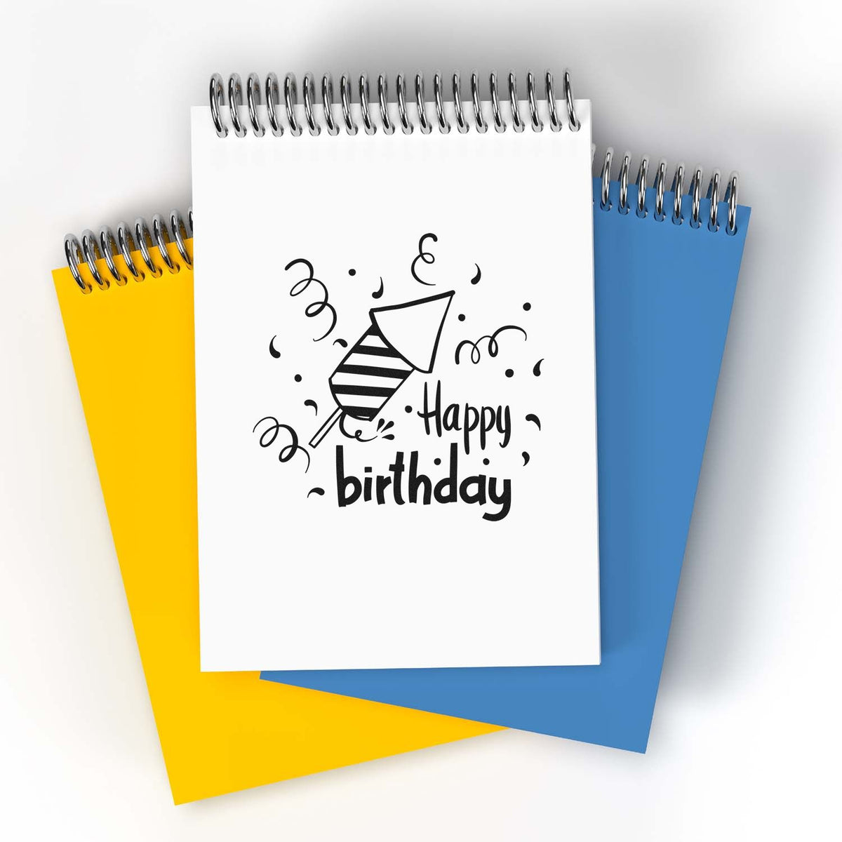 Happy birthday stamp and seal design on white spiral notebook, with black confetti, over yellow and blue notebooks.