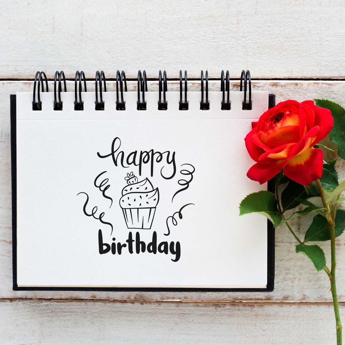 Happy birthday stamp and seal design with black cupcake lettering on white notebook, accented by a red rose.
